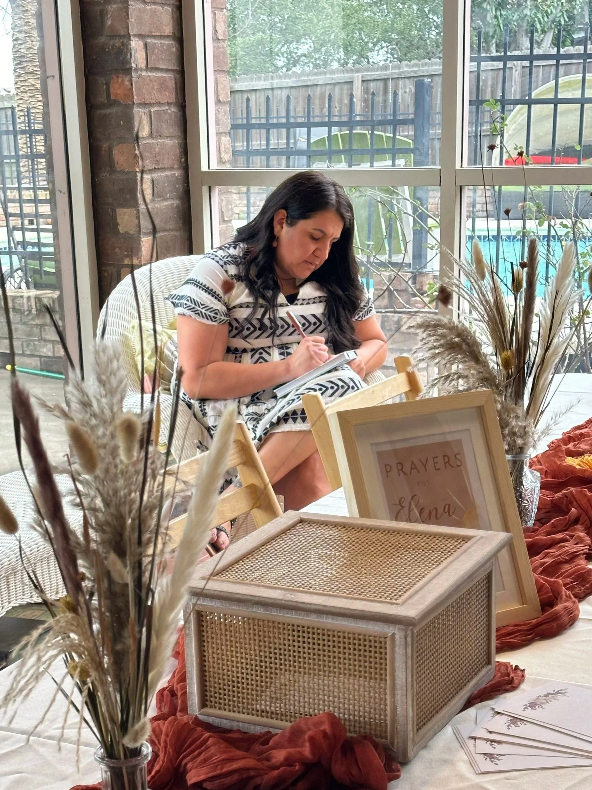 A woman with long dark hair, wearing a black and white patterned shirt, sitting indoors by a large window, writing in a notebook. In the foreground, there are decorative dried plants in a glass jar, and a framed sign that says 'PRAYERS for Elena.' There's a small wooden tray and some papers on the table, and colorful textured cloths around, with a view of a backyard with a fence and outdoor furniture outside the window.