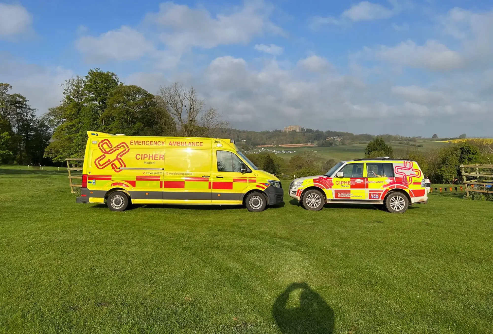 An ambulance and 4x4 car with bright yellow and red colouring face each other in a field, CIPHER is logo'd on their sides in red