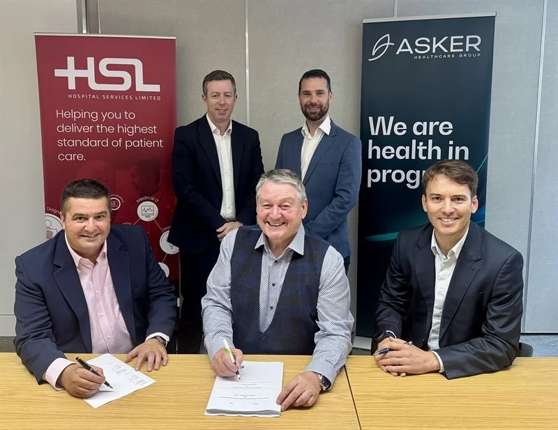 Five male representatives seated and standing around a table signing documents, with HSL Hospital Services Limited and Asker Healthcare Group branded banners behind them.