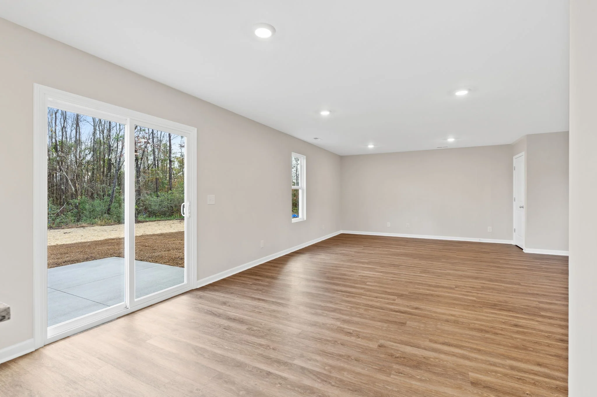 Empty living room with light-colored walls, wooden floor, sliding glass door to outdoor patio, and small windows, illuminated by ceiling lights.