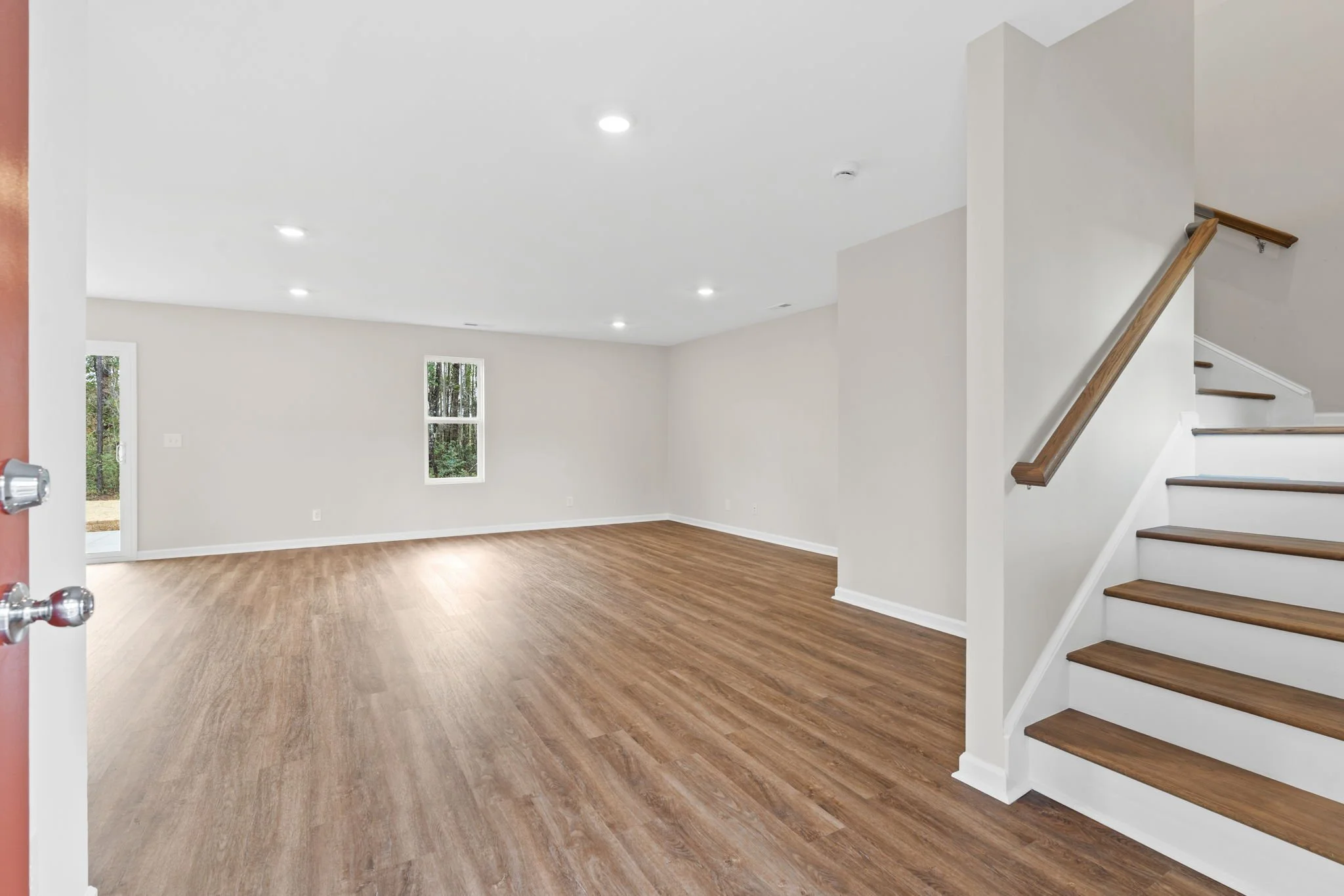 Empty living room with wood flooring, white walls, two windows, and a staircase with wooden handrail on the right.