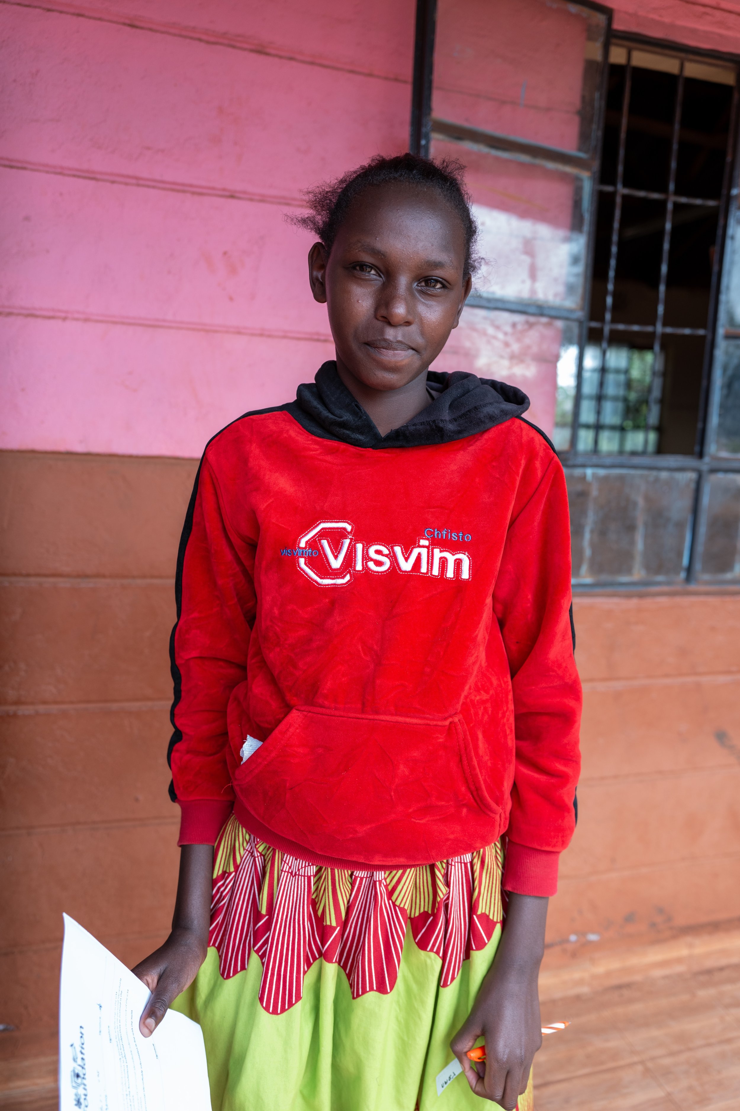 A young girl standing in front of a wooden wall, wearing a red hoodie with white and blue embroidery and a colorful skirt, holding a piece of paper in her right hand.