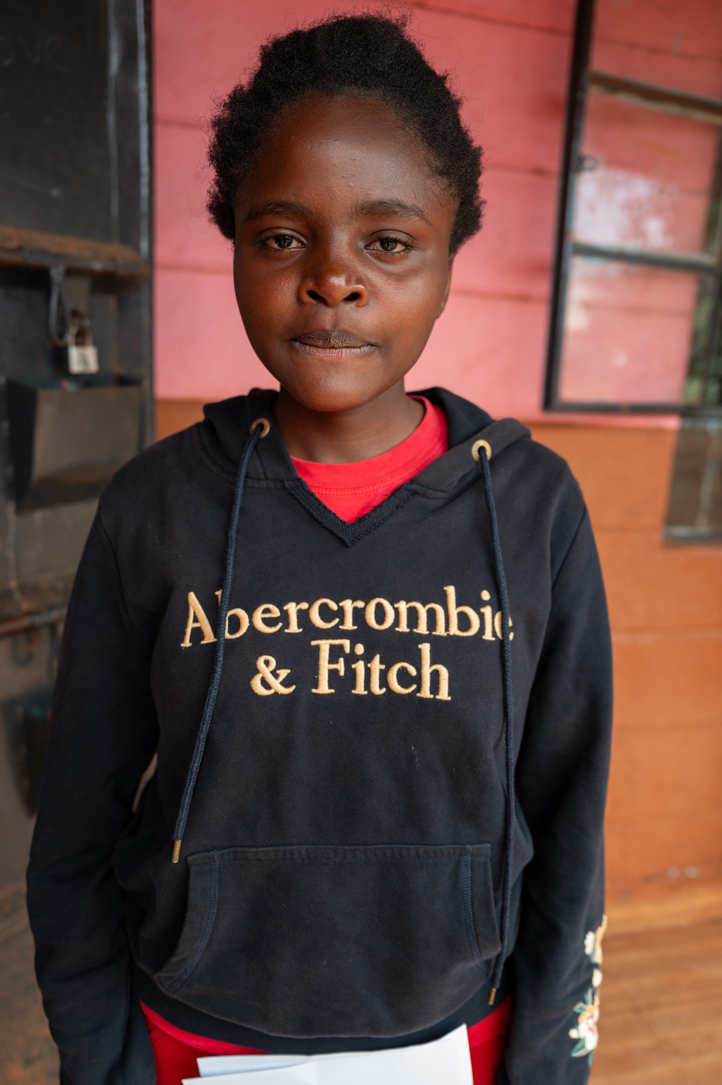 A young girl standing indoors, wearing a black Abercrombie & Fitch hoodie with a red shirt underneath, and holding a piece of paper.
