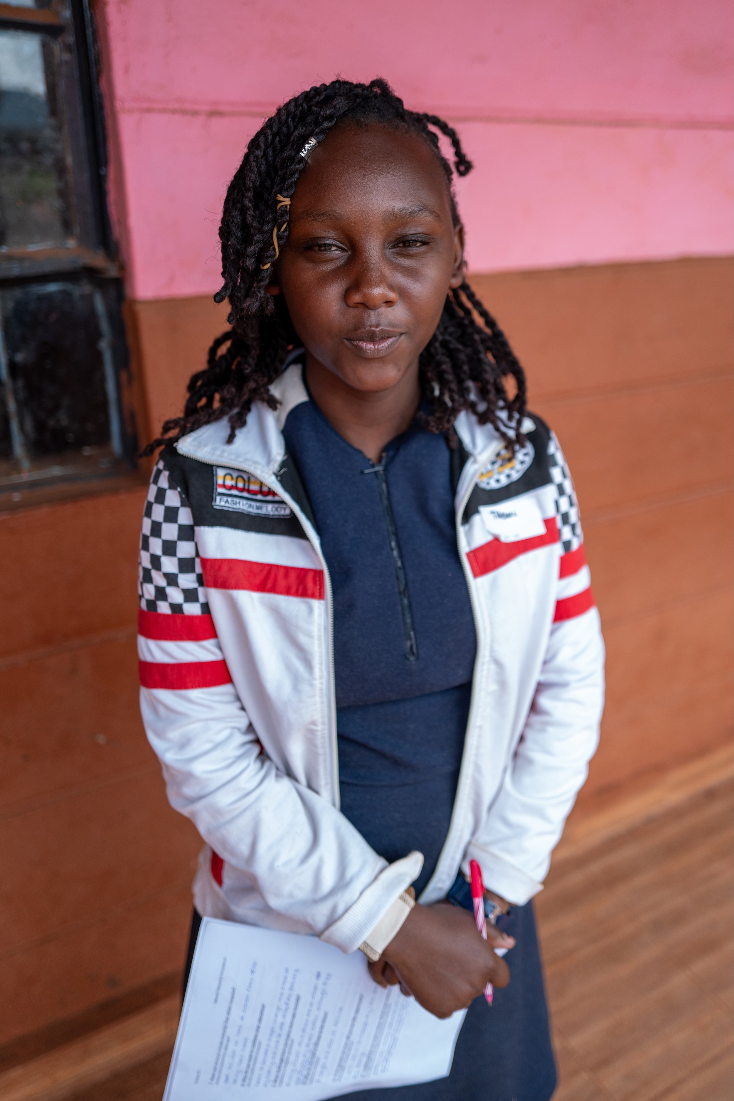 A young girl with braided hair, wearing a navy blue top and a white jacket with red and black checkered pattern, holding a pen and paper, standing against a wooden and pink wall.