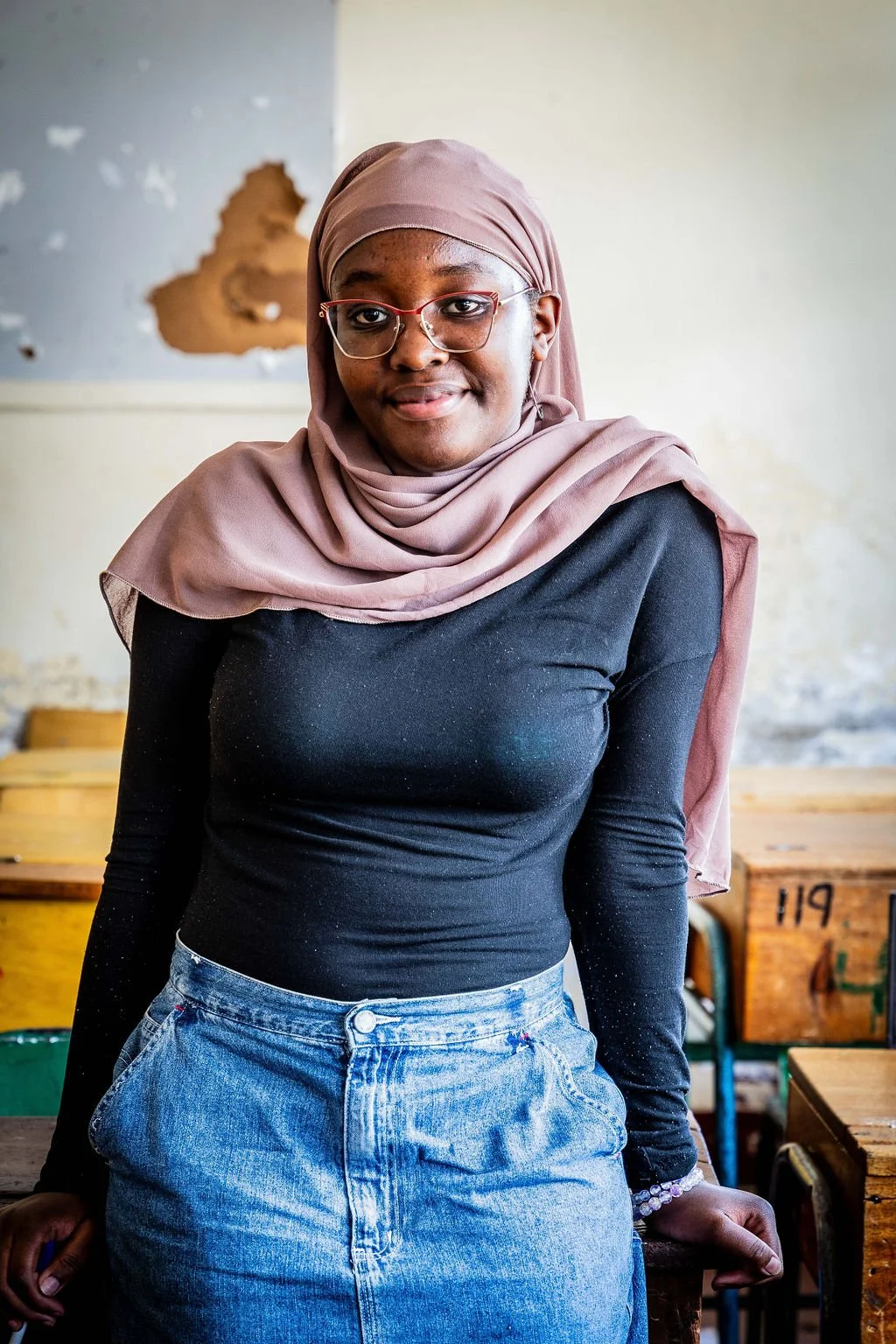 Person wearing glasses and a hijab, standing in a classroom.