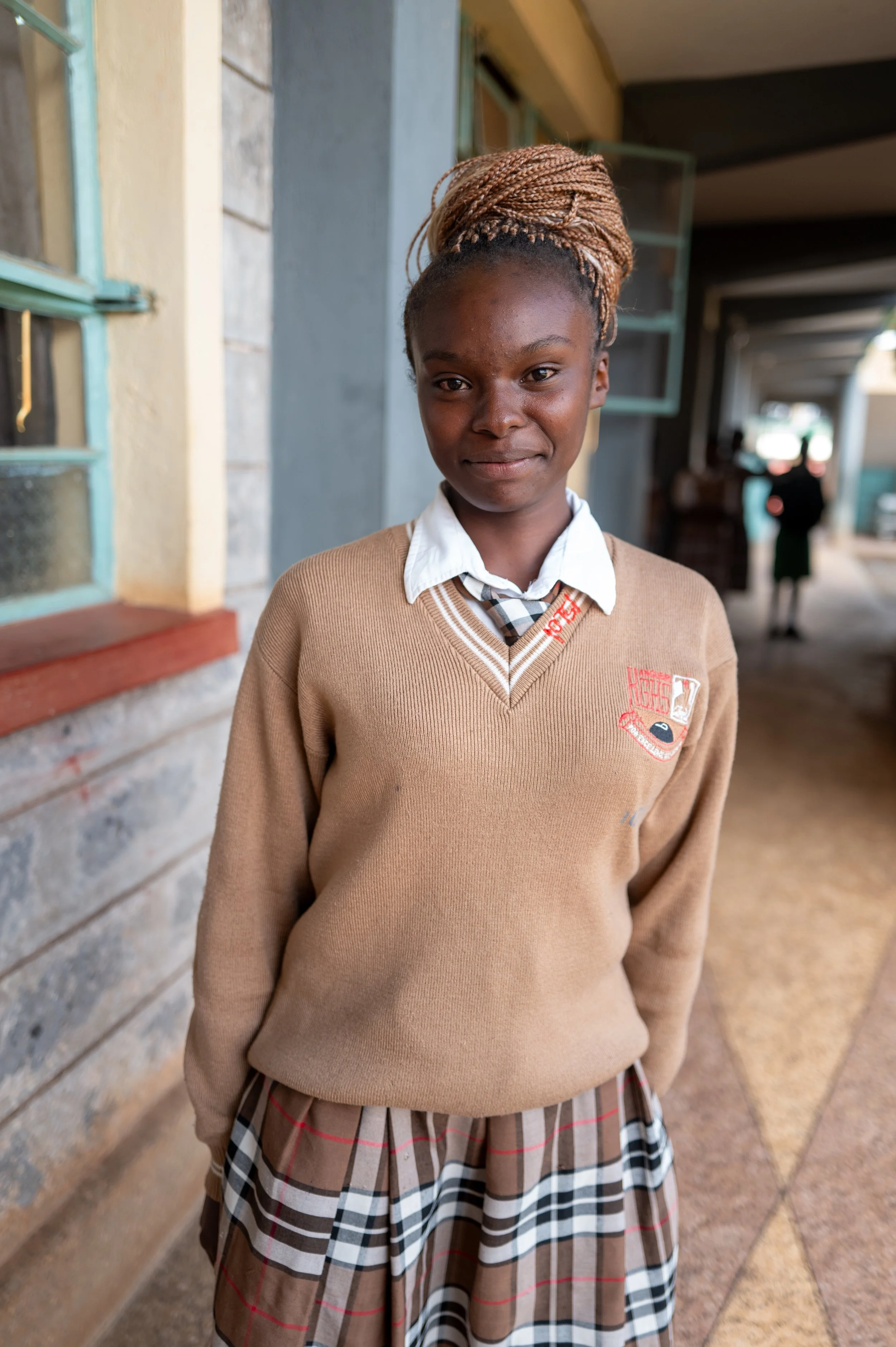 A young girl in a school uniform standing outside a building, with a slight smile on her face.