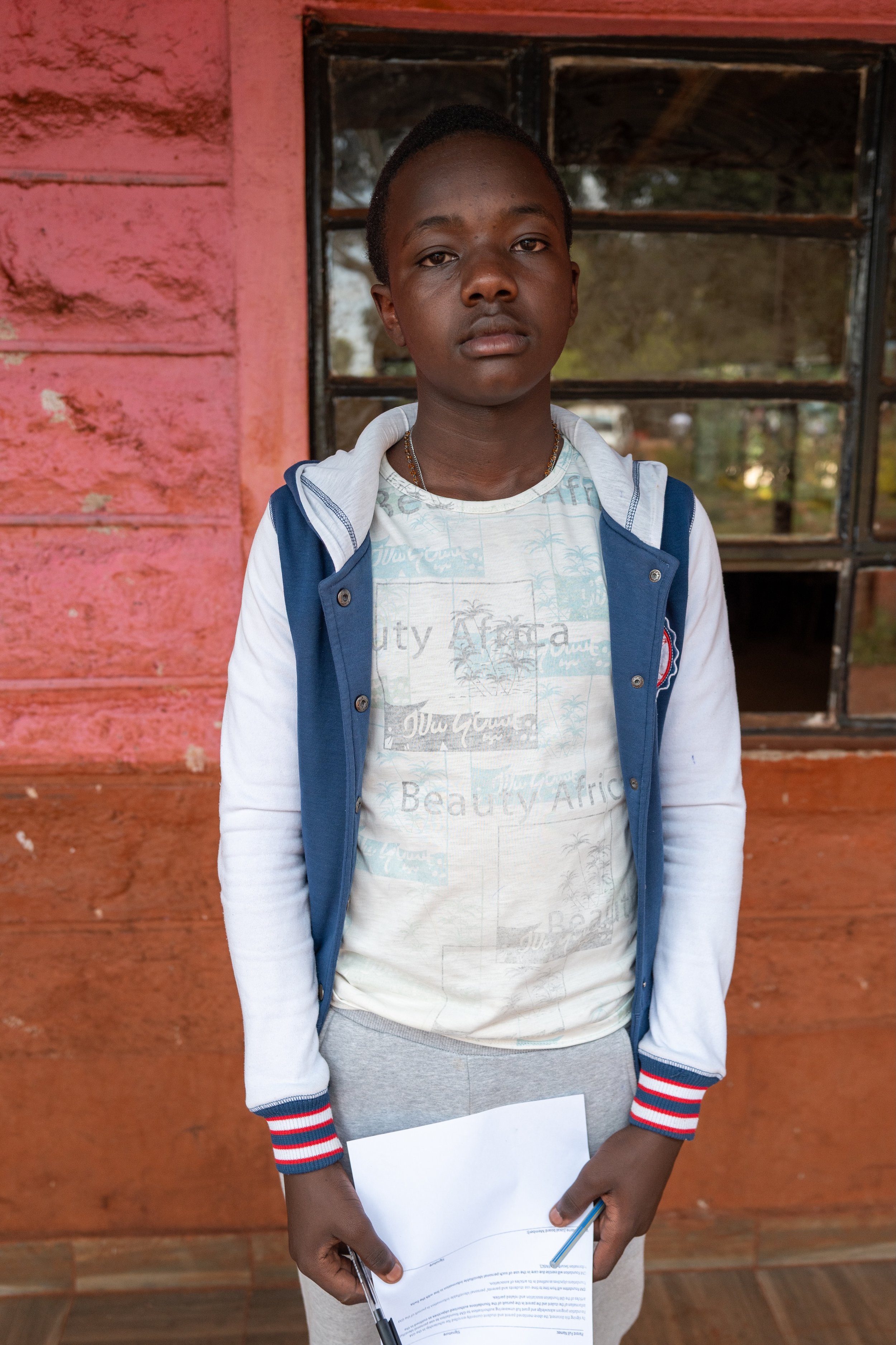 A young boy stands in front of a red and black windowed wall, wearing a white shirt with light blue patterns, a blue and white jacket, and grey sweatpants. He holds a paper and pen in his hands, looking directly at the camera with a serious expressio
