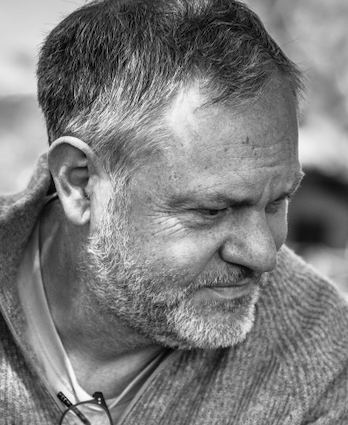 A man with gray hair and a beard wearing a blue blazer and black shirt, posing against a light background.