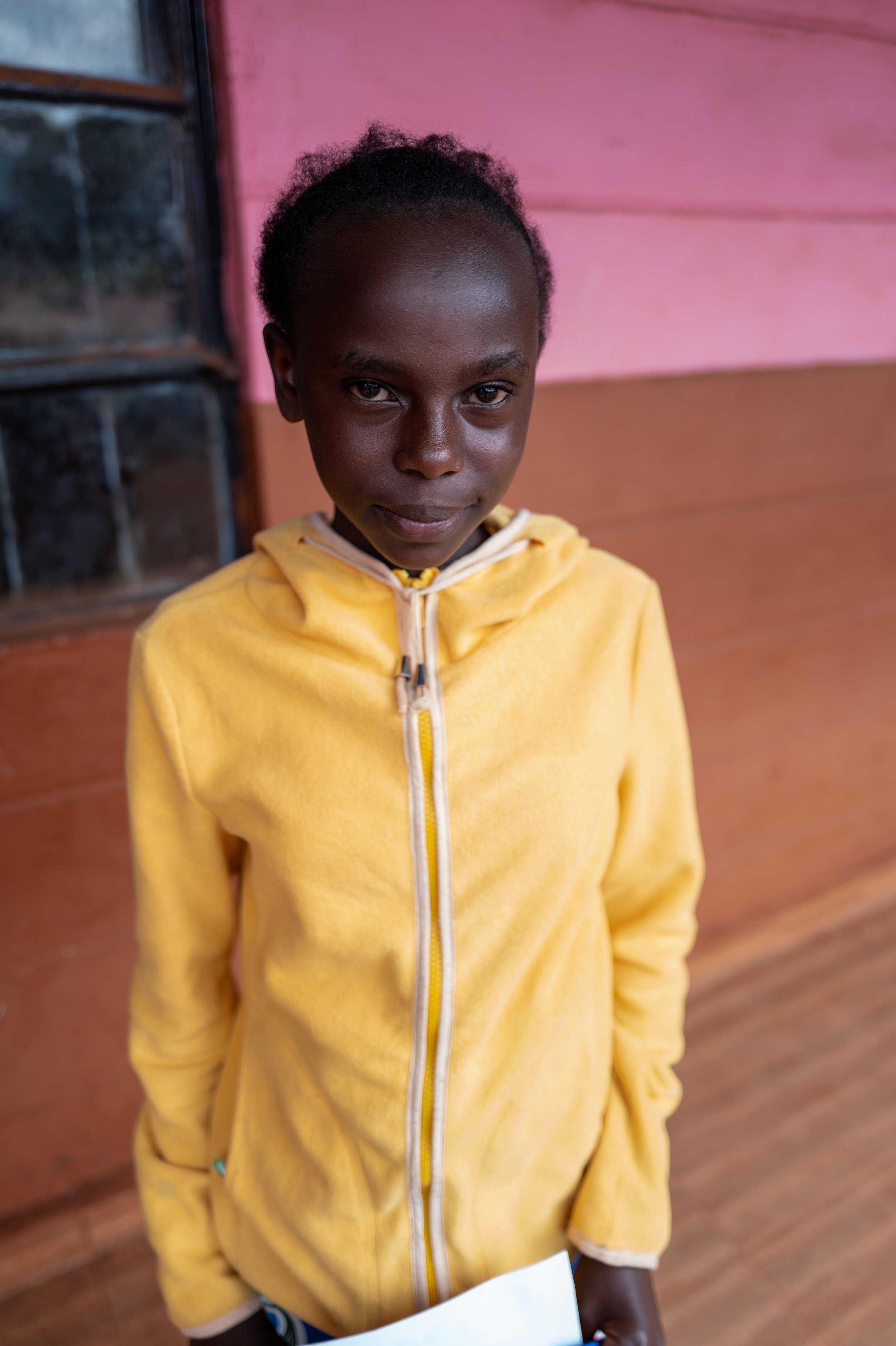A young girl wearing a yellow hoodie is standing indoors against a colorful wooden wall, looking at the camera.