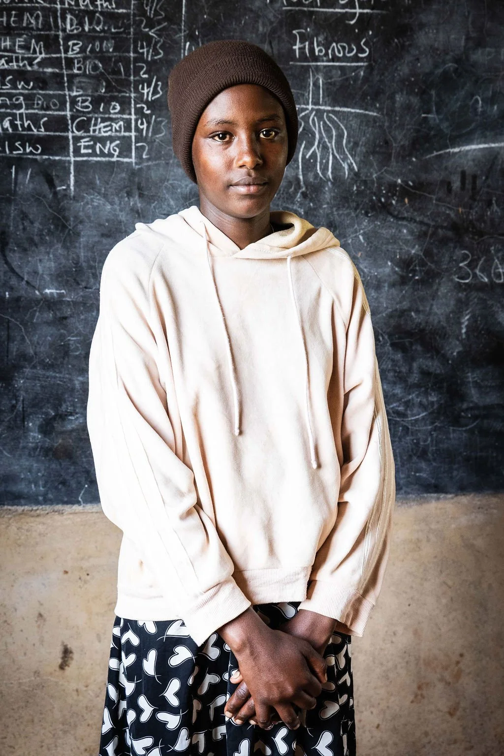 Person wearing a hoodie and hat stands in front of a chalkboard with writing.