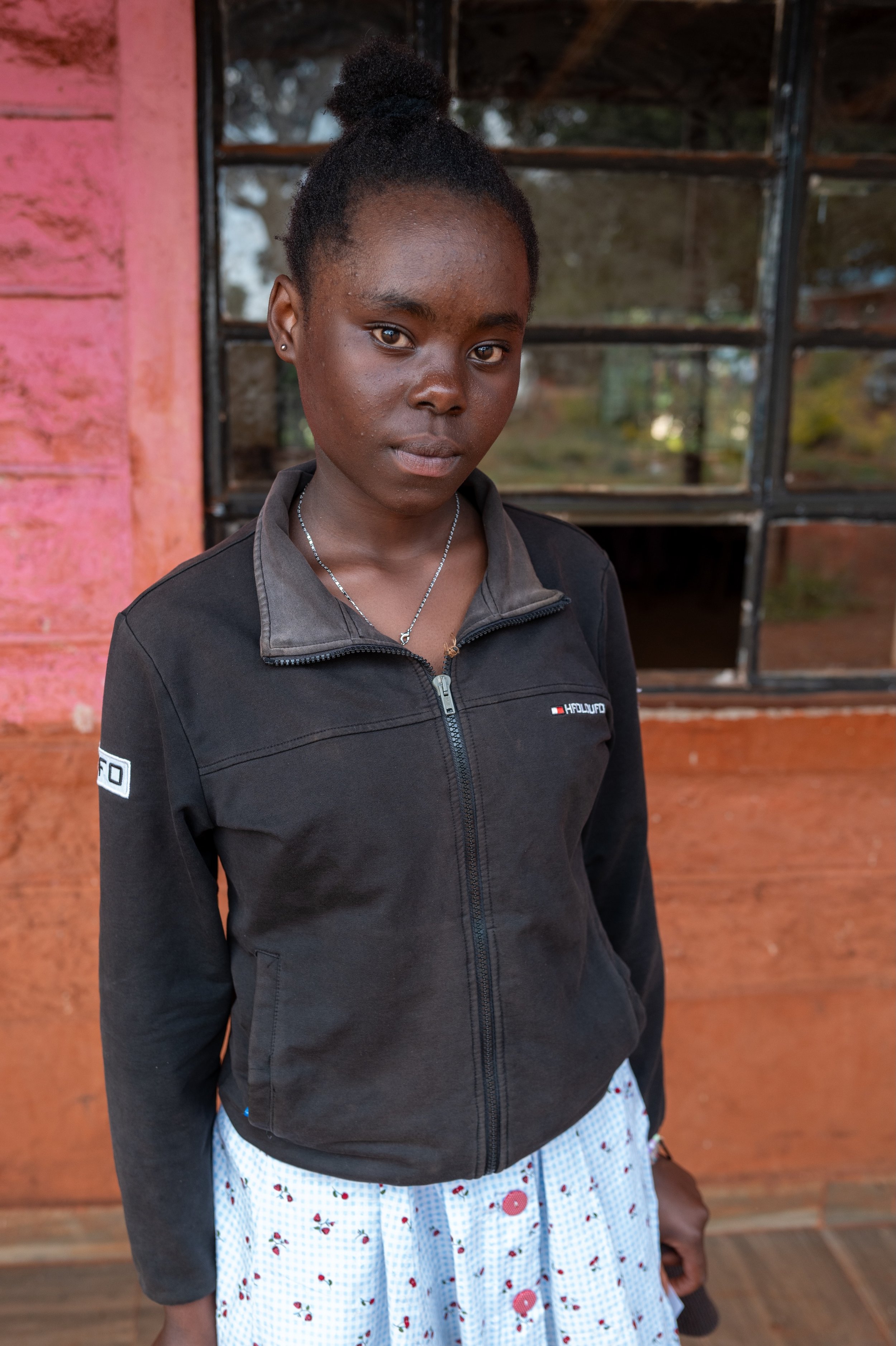 A young woman with dark skin and curly hair tied up, wearing a black zip-up jacket and a white skirt with red and blue pattern, standing outside in front of a rustic, pink and brown wall with window panes.