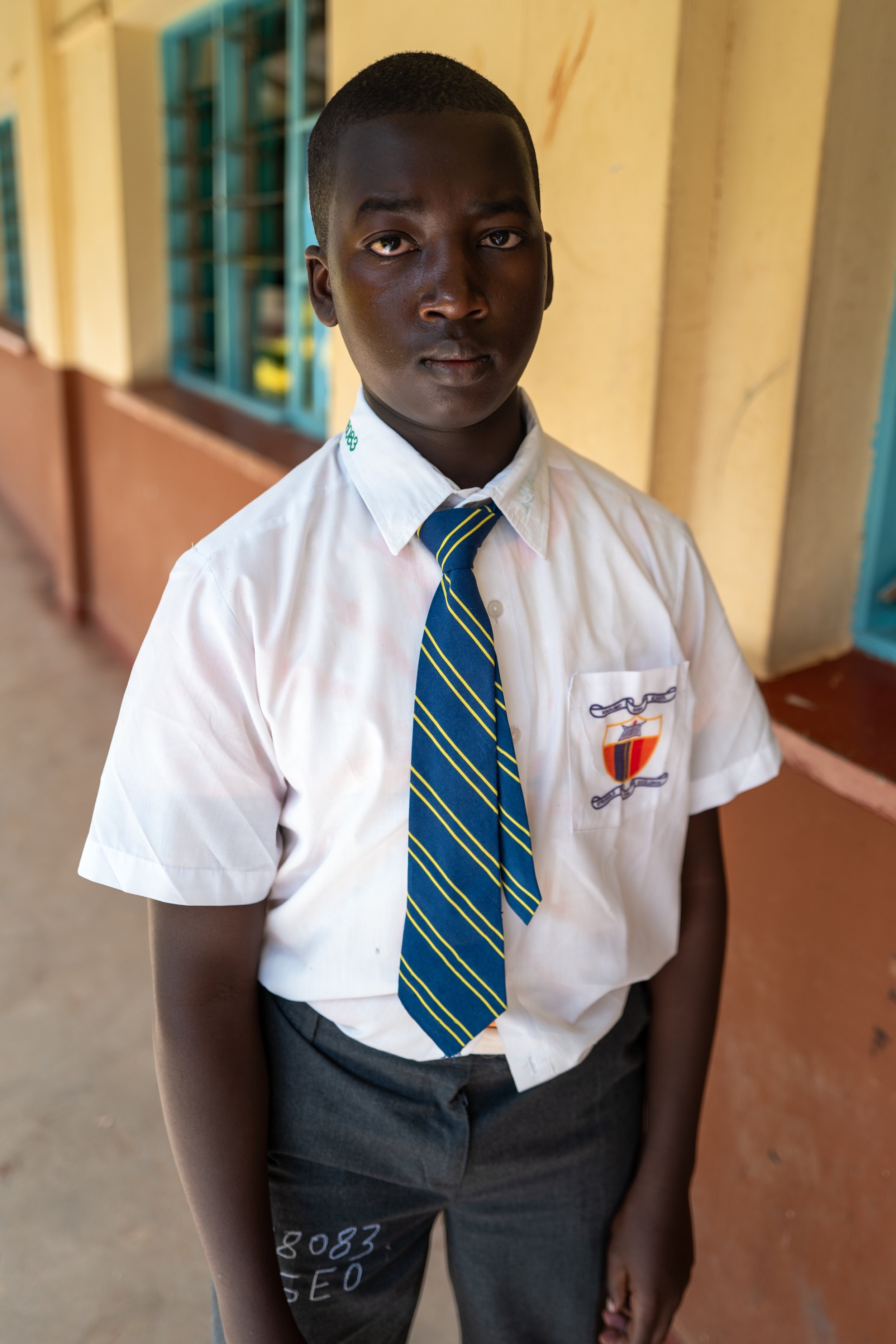 A young African boy in a school uniform standing in a school corridor, looking at the camera.