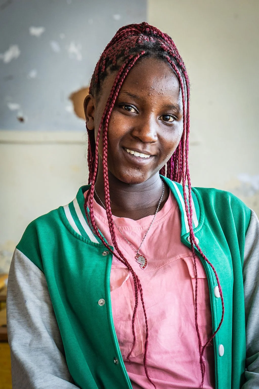 A person smiling at the camera, with red braided hair and wearing a green and gray jacket over a pink shirt. The background is a light-colored wall.