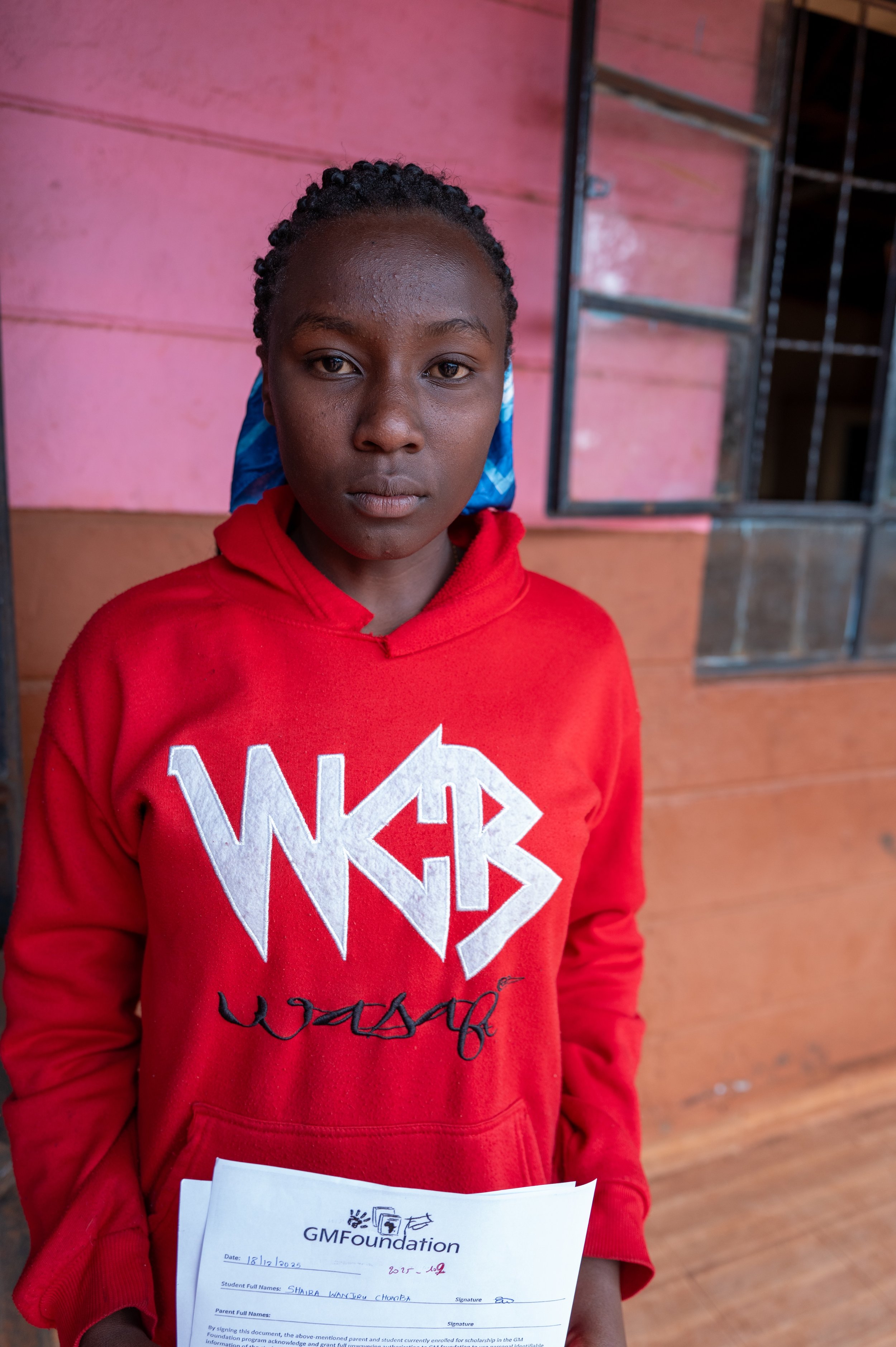 A girl standing inside a building with pink walls, holding a document, wearing a red hoodie with white and black lettering, and a blue headscarf.