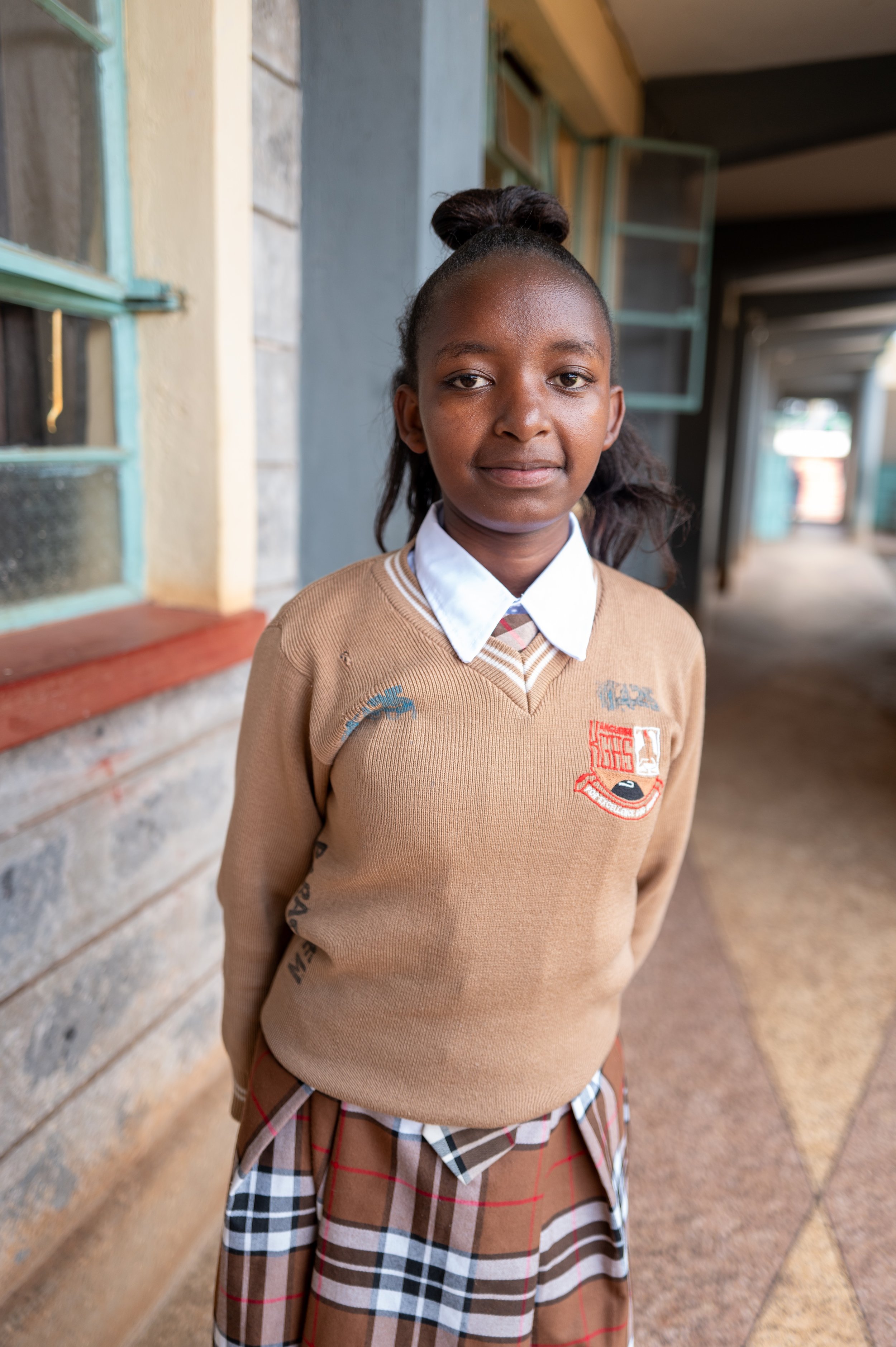A young girl in a school uniform stands outdoors beside a building with brick and plaster walls, under an open corridor, and looks at the camera with a gentle smile.