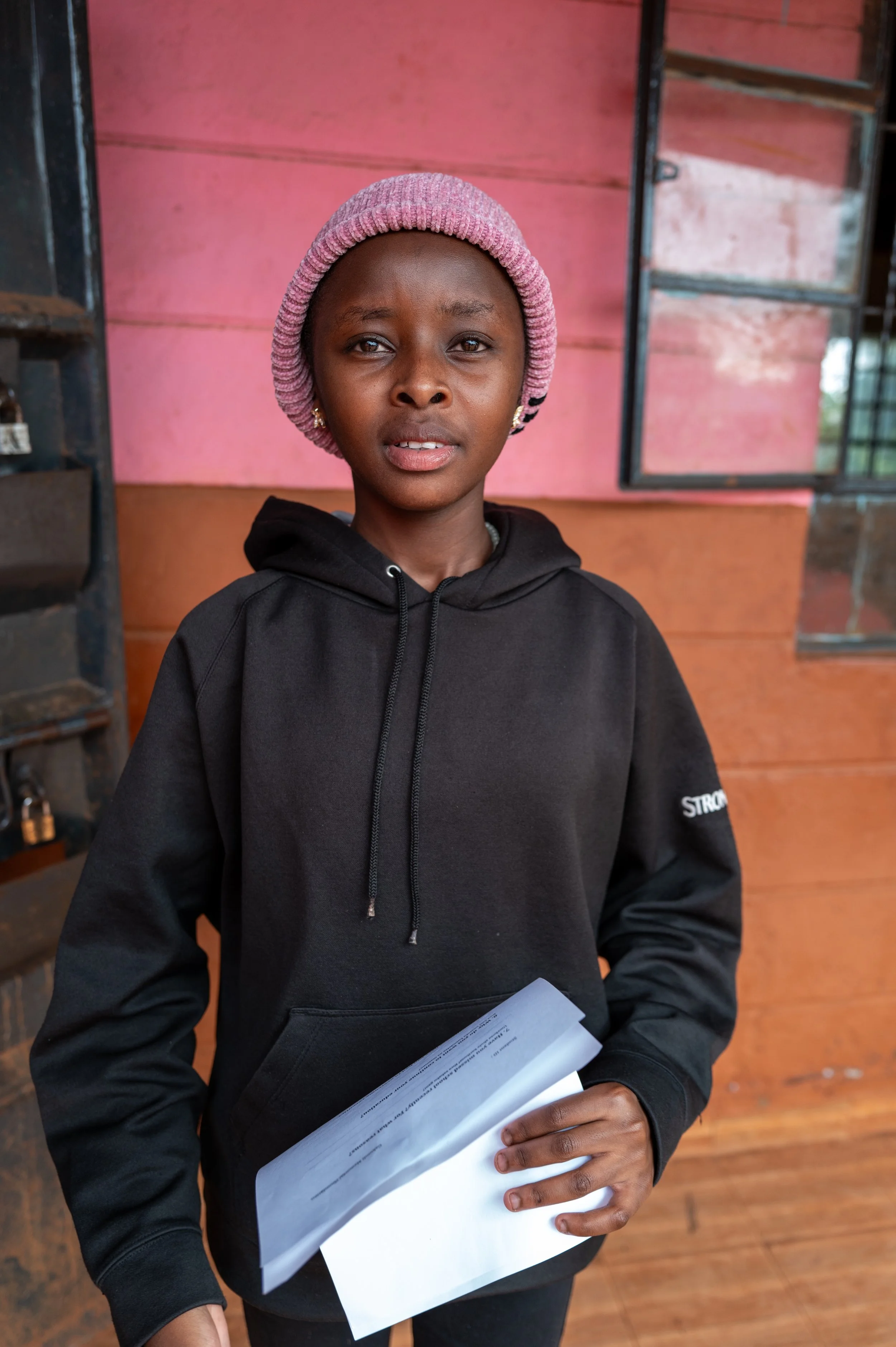 Young girl wearing a pink knit cap and black hoodie, holding papers, standing indoors with pink and brown wooden wall behind her.