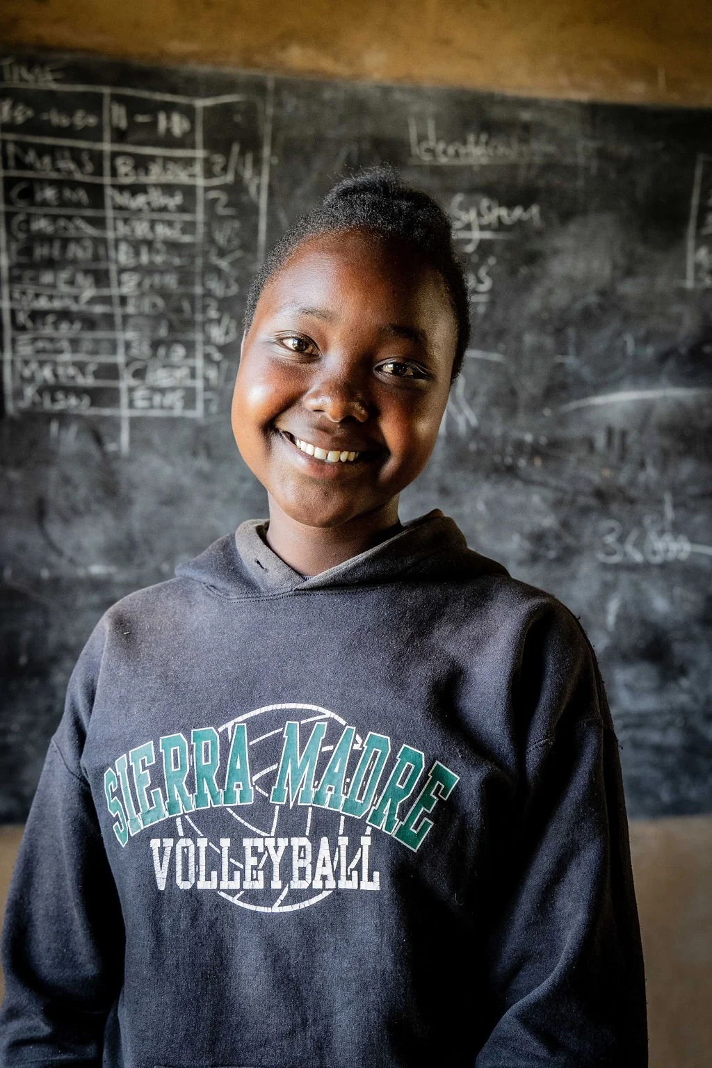 A smiling student wearing a "Sierra Madre Volleyball" sweatshirt stands in front of a chalkboard with writing on it.