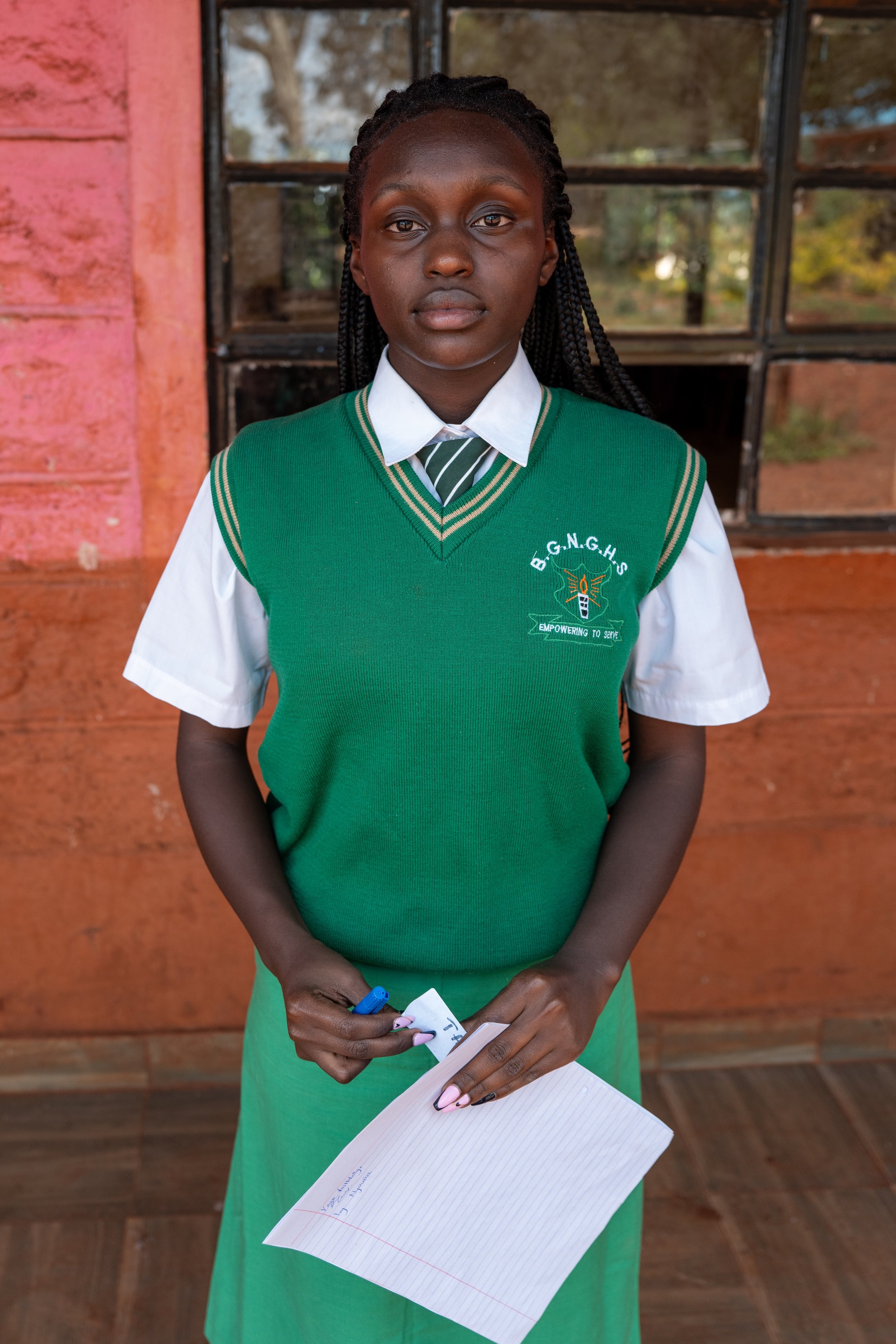 A young girl in a school uniform standing indoors, holding a notebook and a blue pen. She is wearing a white shirt, a green vest with a school logo, and a striped tie. Behind her is a window with a view of trees outside.