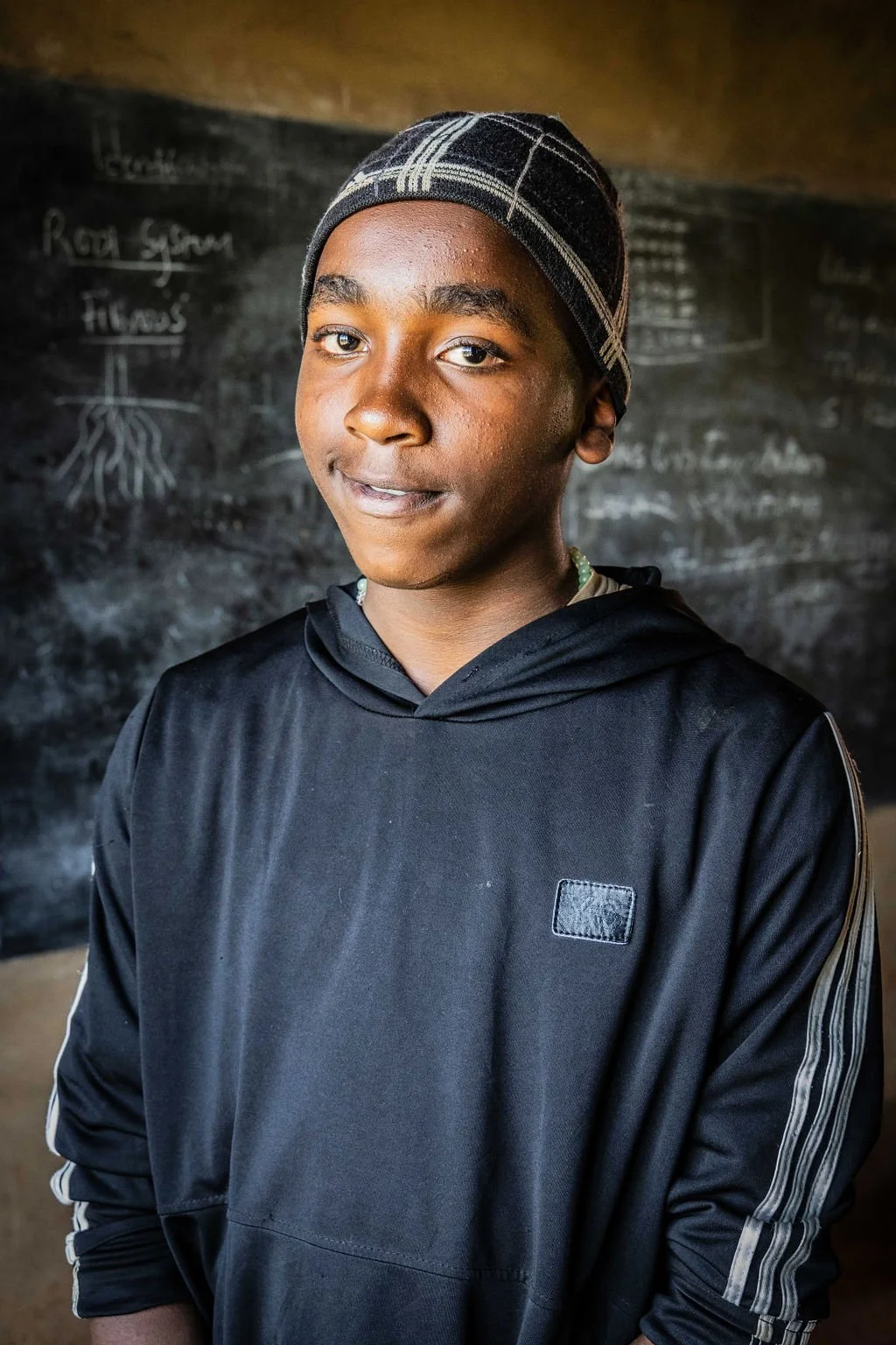 Young person wearing a black hoodie and beanie, standing in front of a chalkboard.