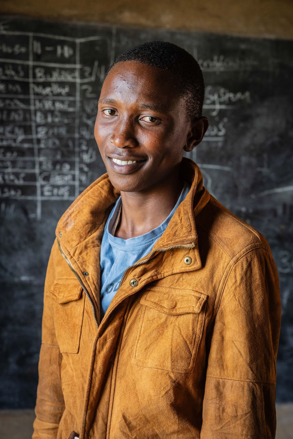 Person in brown jacket smiling in front of a chalkboard with writing.