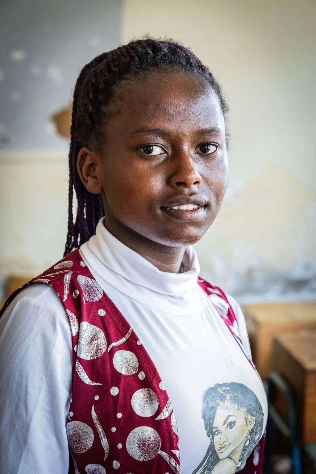 A young person with braided hair is wearing a white turtleneck and a red patterned vest, standing indoors.