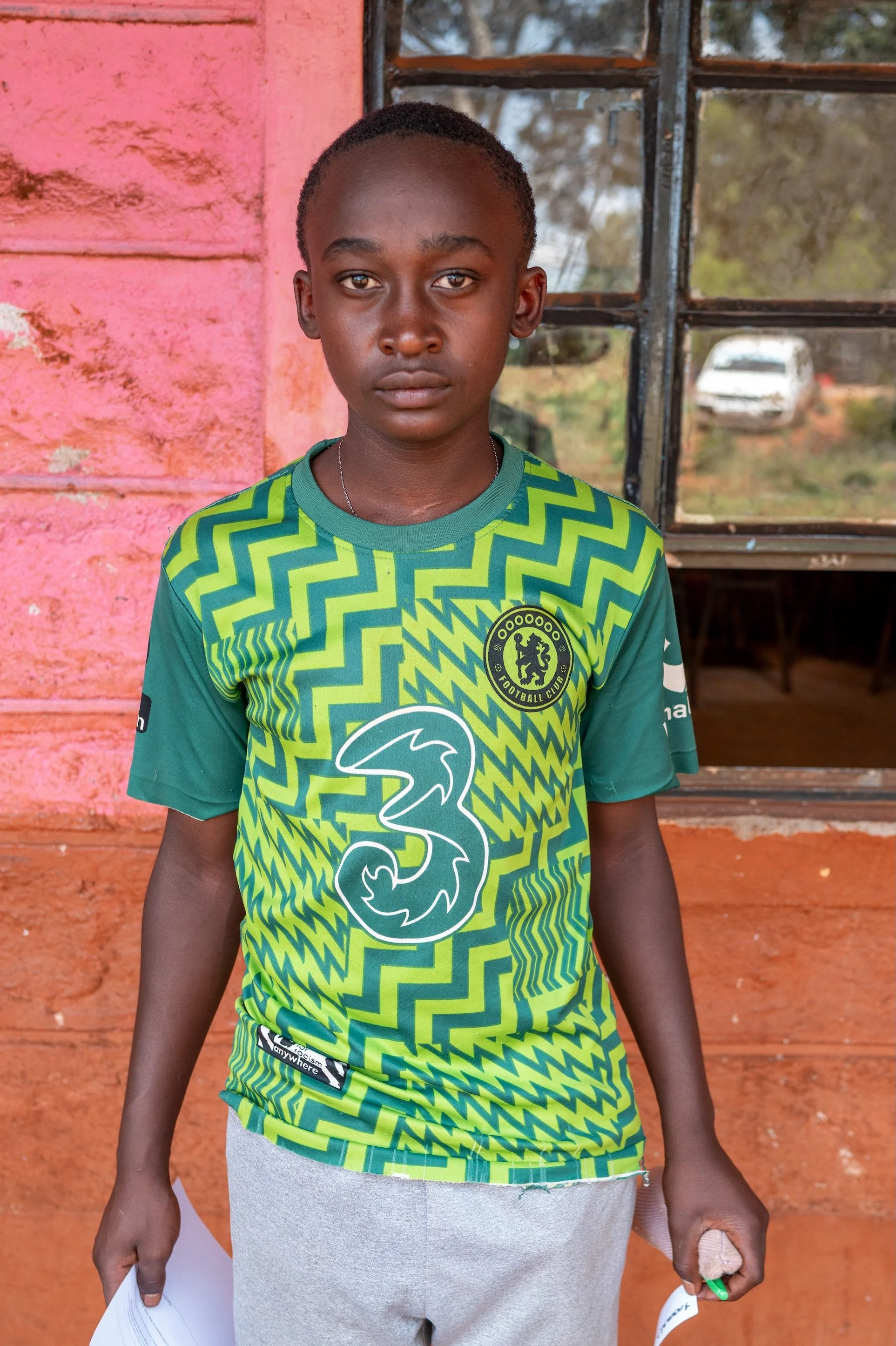 A young boy stands outside a building with pink and brick walls, wearing a green Chelsea football jersey, holding papers and a green pen.