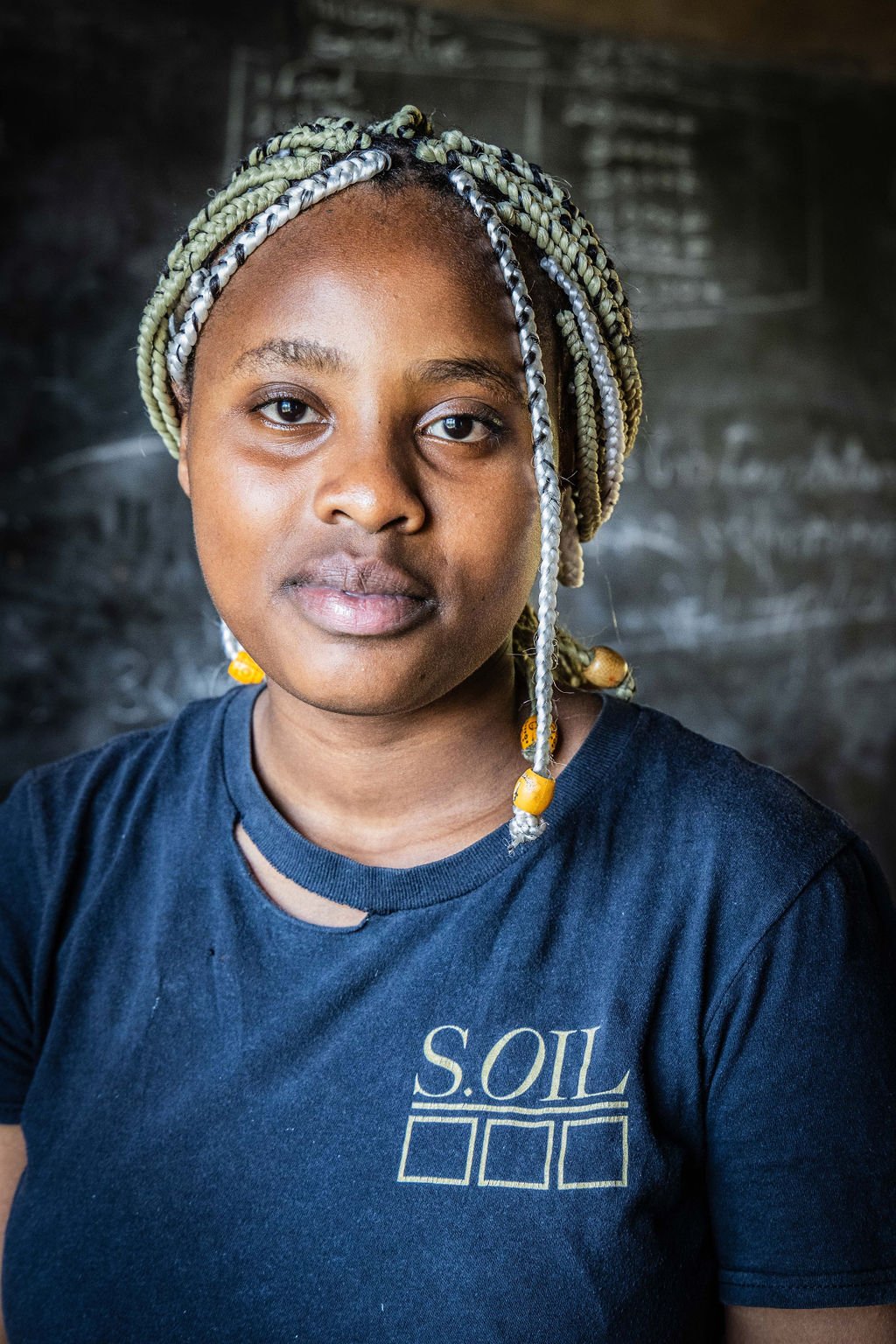Person with braided hairstyle wearing a blue shirt with S.OIL logo, standing in front of a chalkboard.