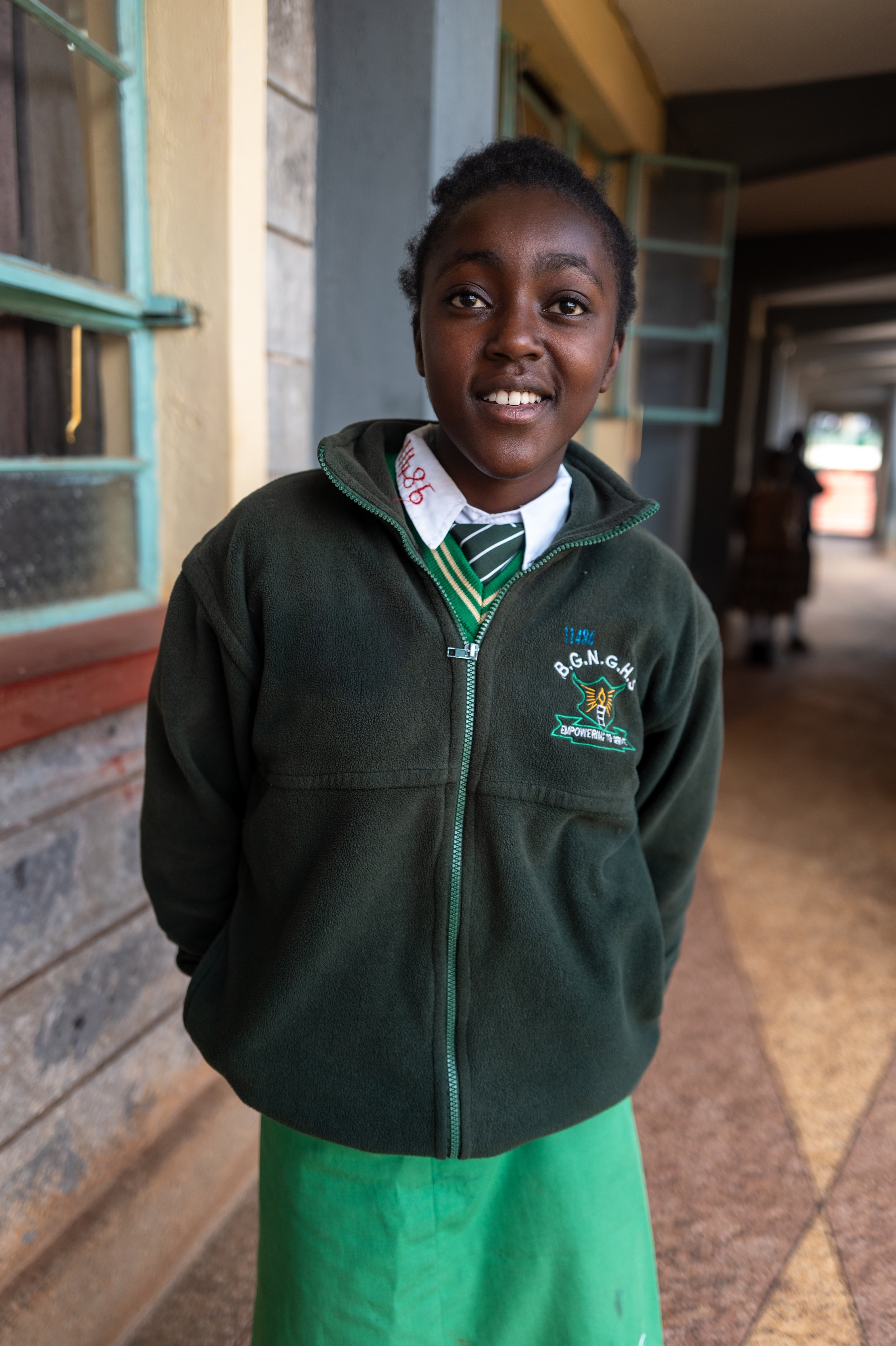 A young girl in a school uniform standing inside a building near a window, smiling at the camera.