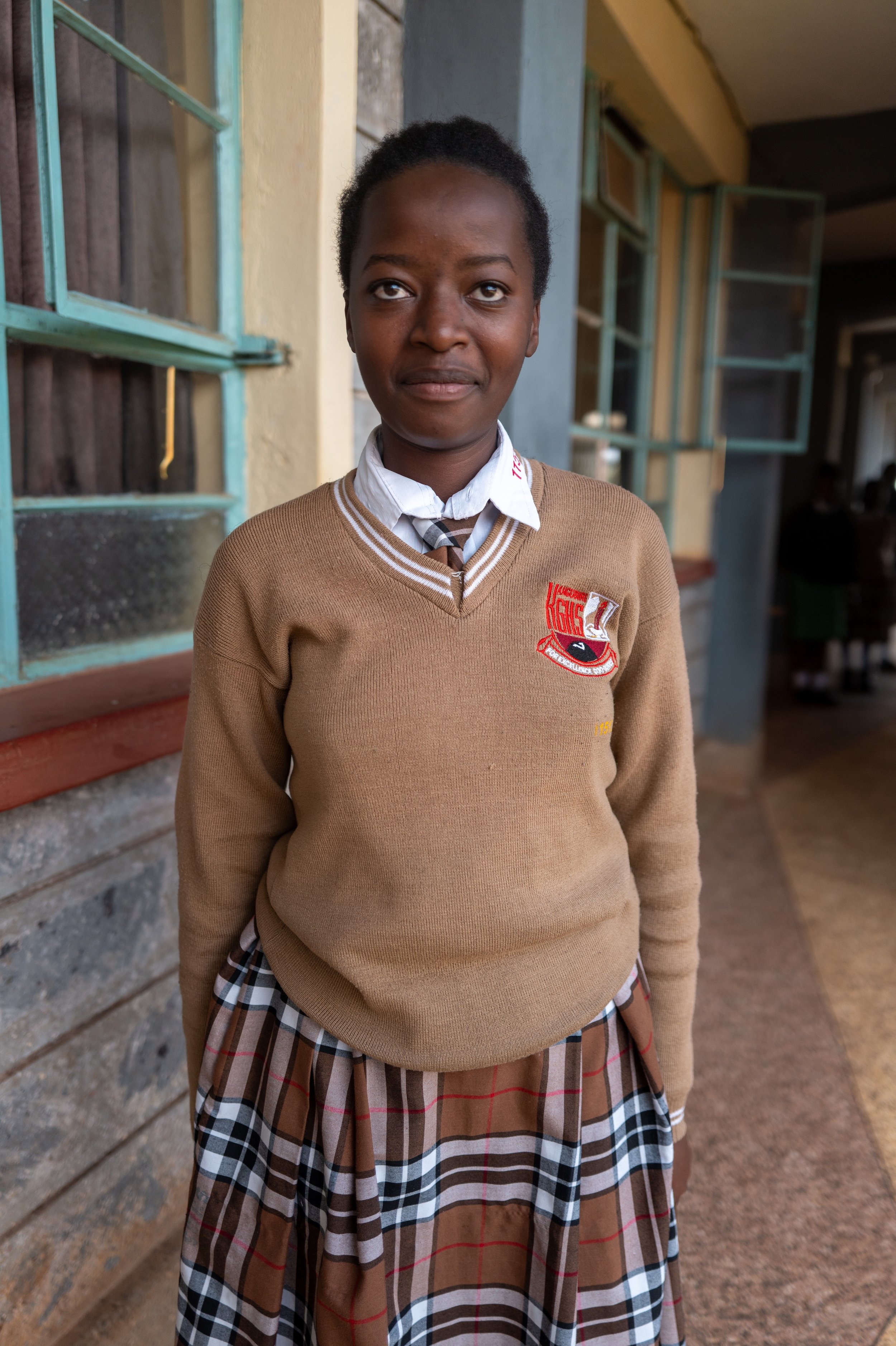 A young girl in a school uniform standing outside a building, with open windows behind her.