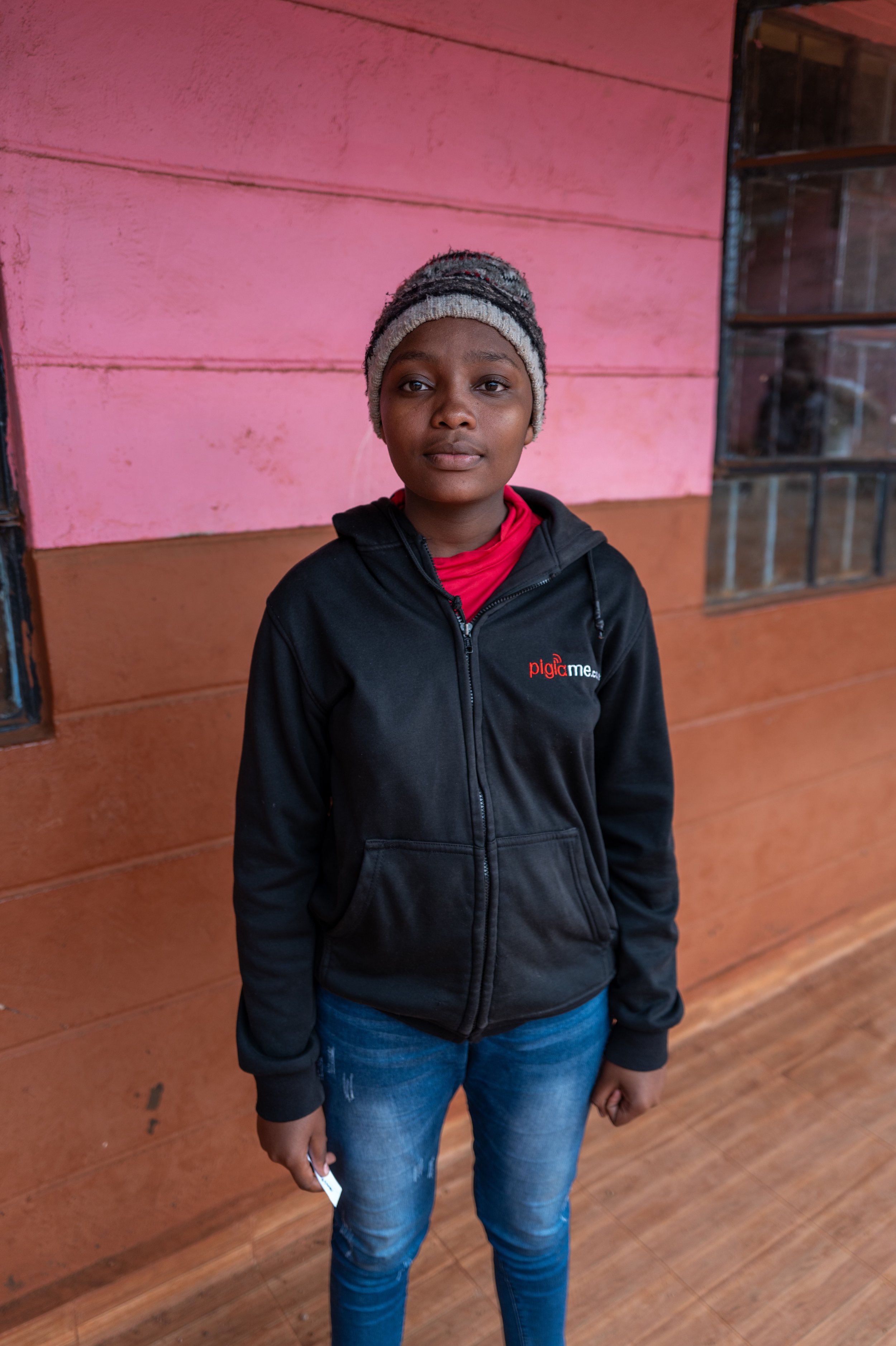 A young girl wearing a knit hat and a black hoodie with red and white text, standing indoors in front of a pink and orange wooden wall.