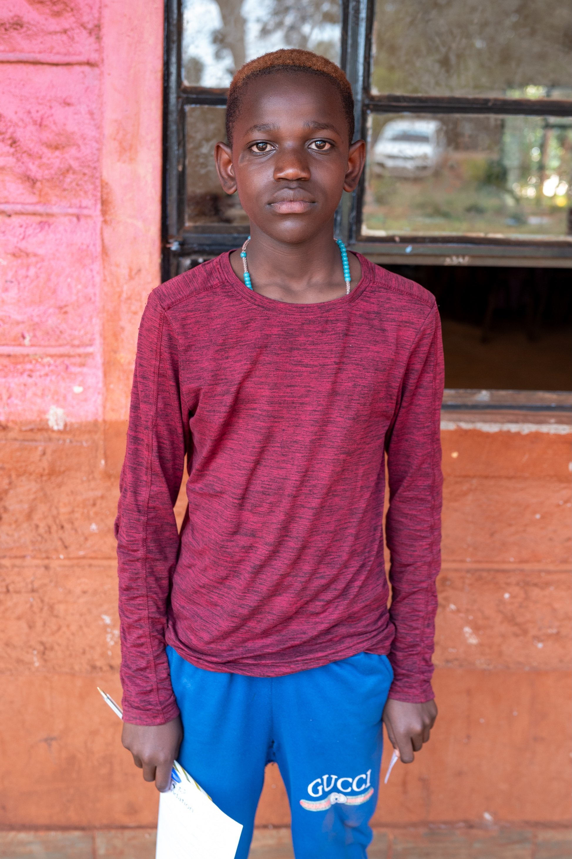 A young boy standing indoors in front of a painted wall and a window, wearing a red long-sleeved shirt, blue pants with 'GUCCI' written on them, and holding papers. He has a short hairstyle and is wearing a beaded necklace.