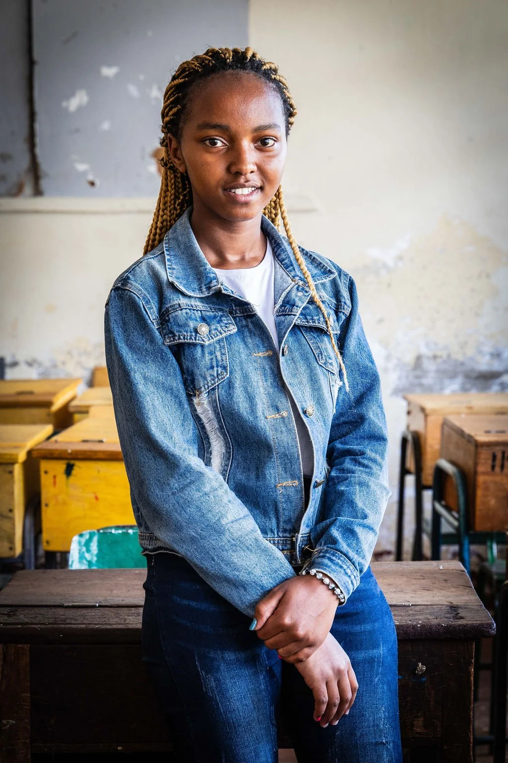 Young woman with braided hair wearing a denim jacket and jeans, standing indoors with old desks in the background.