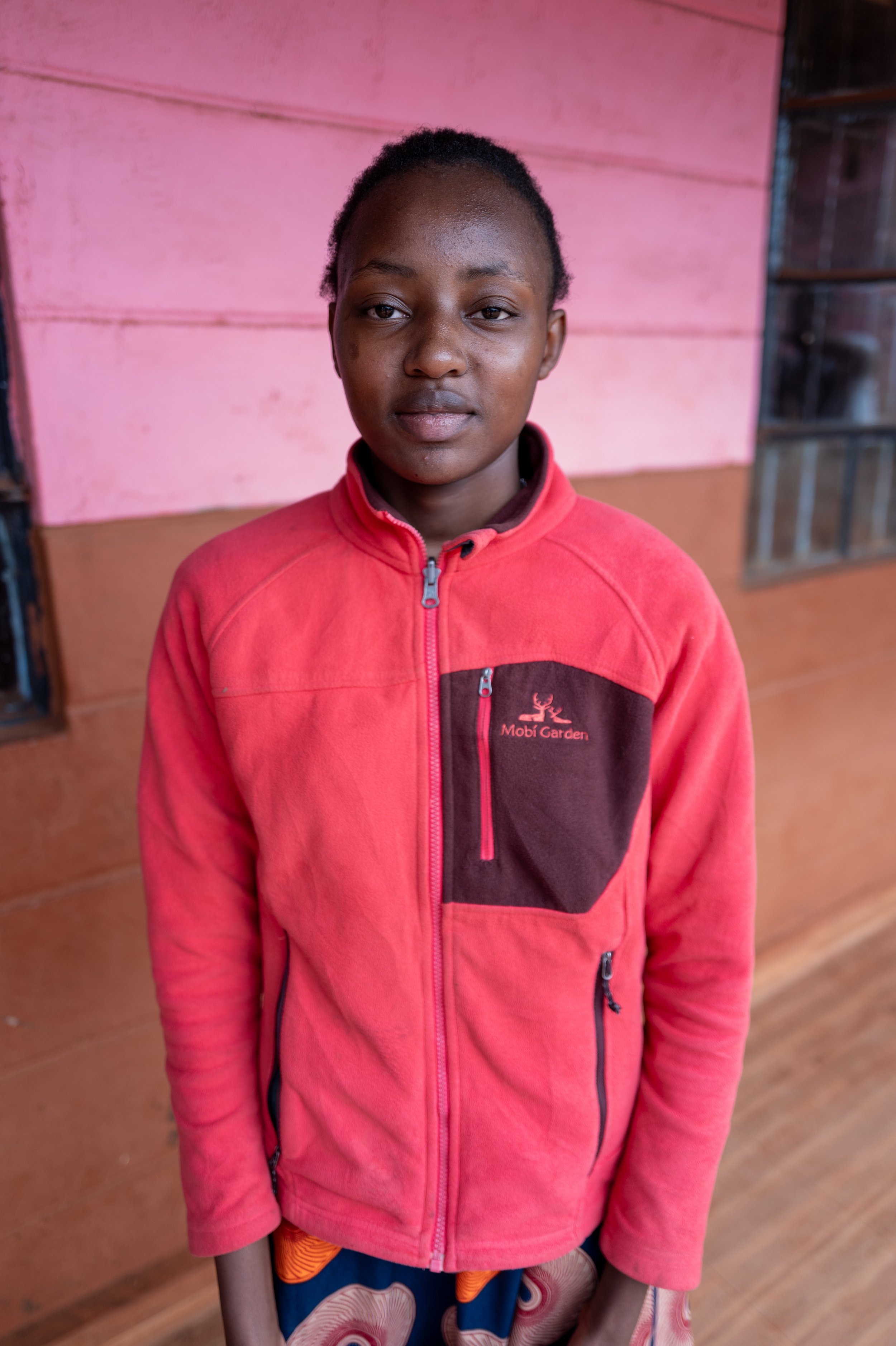 A young woman with short hair wearing a red fleece jacket standing in front of a pink and brown wooden wall.
