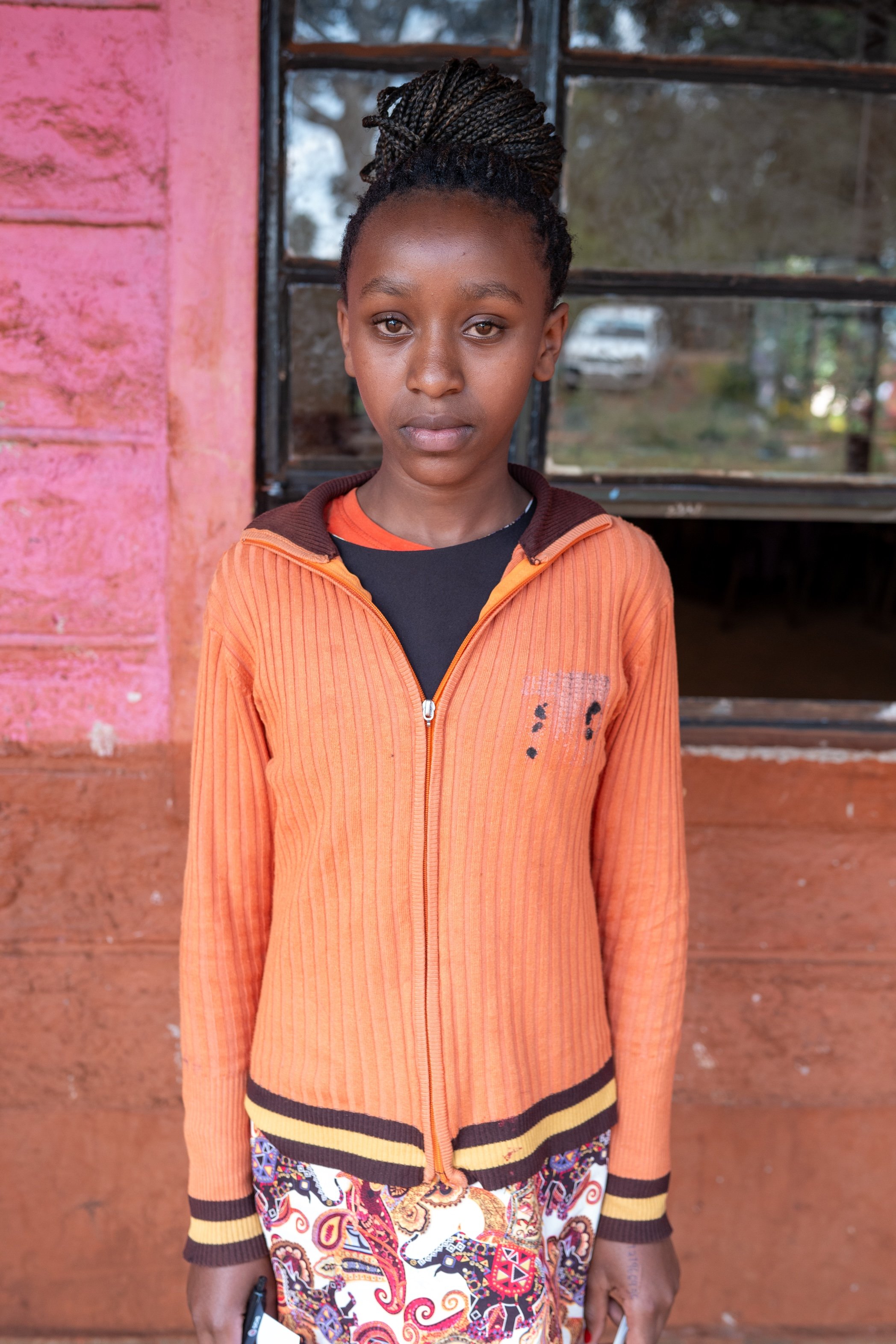A young girl with braided hair standing in front of a pink and brown wall near a window.