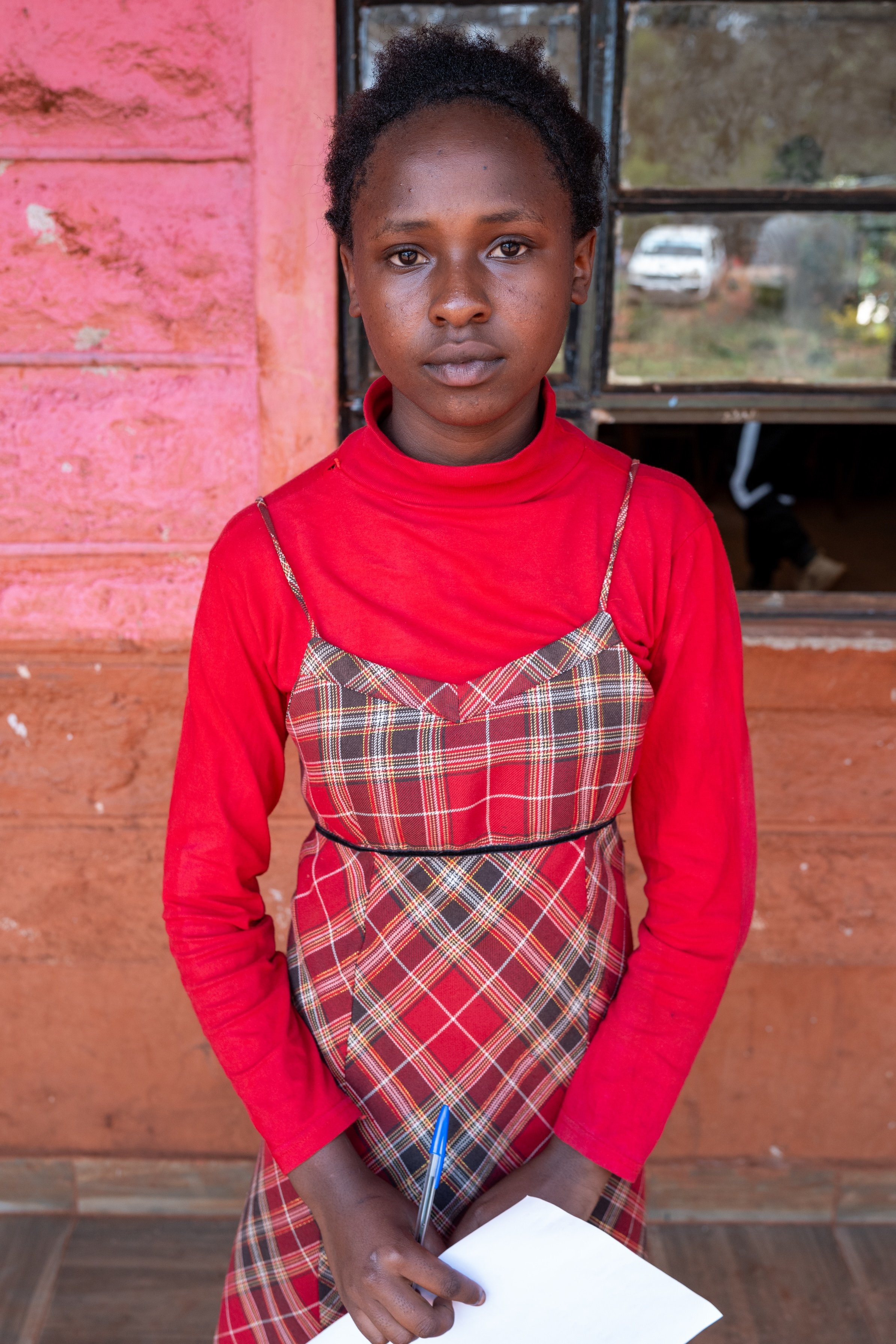 Young girl in a red long-sleeve shirt and plaid jumper holding a pen and paper, standing in front of a pink and brown wall with a window.