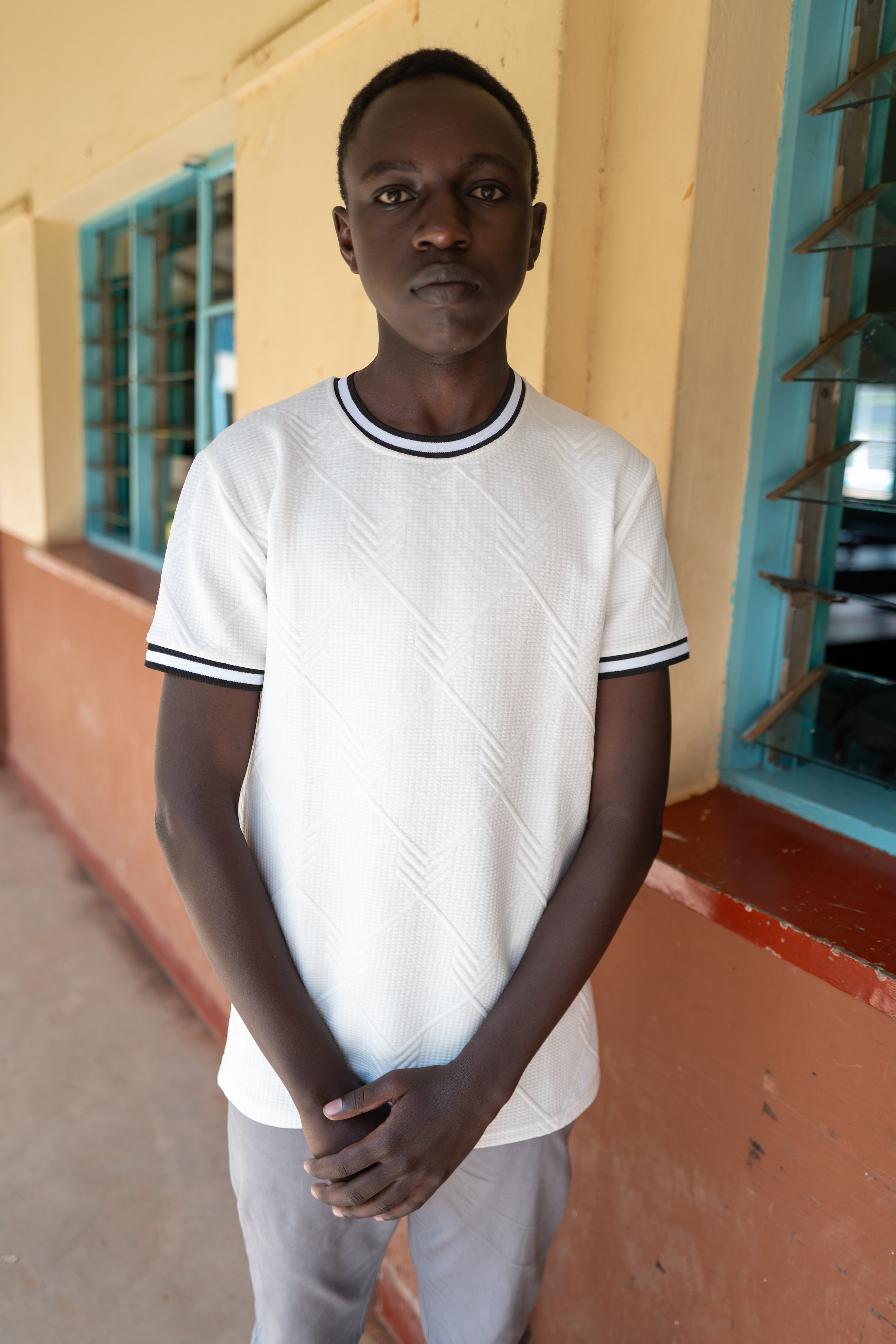 A young man with dark skin and short black hair standing indoors near a window, wearing a white T-shirt with black-striped collar and sleeves, and light gray pants.