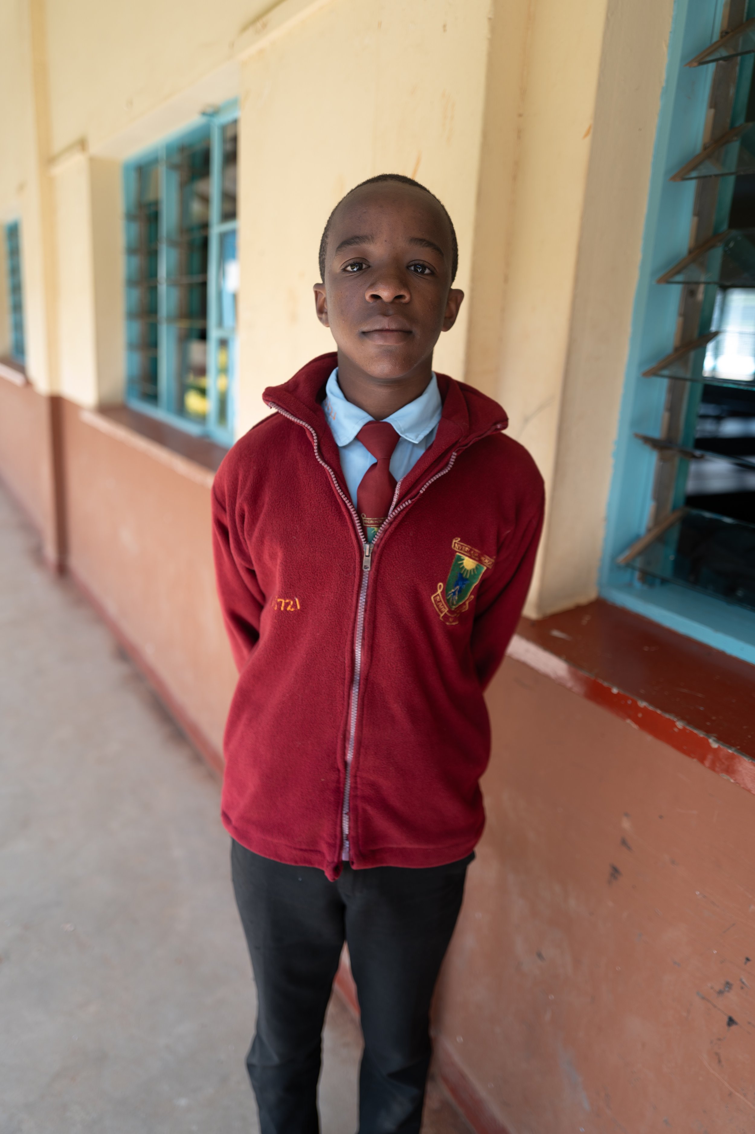 A young boy in a school uniform standing in a hallway with yellow walls and blue window frames, facing the camera.