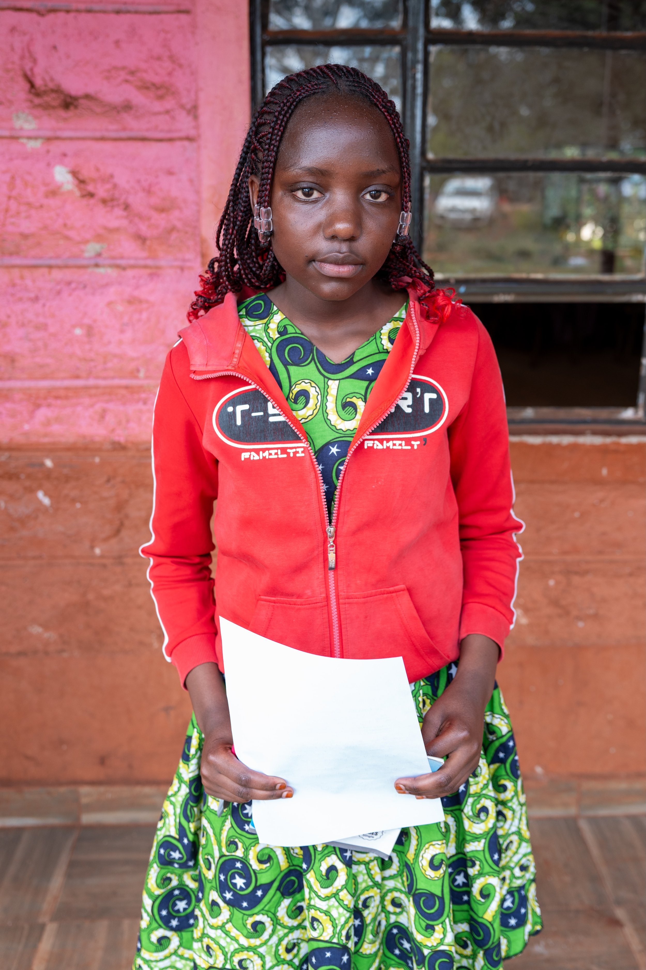 A young girl with braided hair wearing a red jacket with a patterned green dress underneath is holding a piece of white paper standing inside a building with a pink and brown wall and a window behind her.