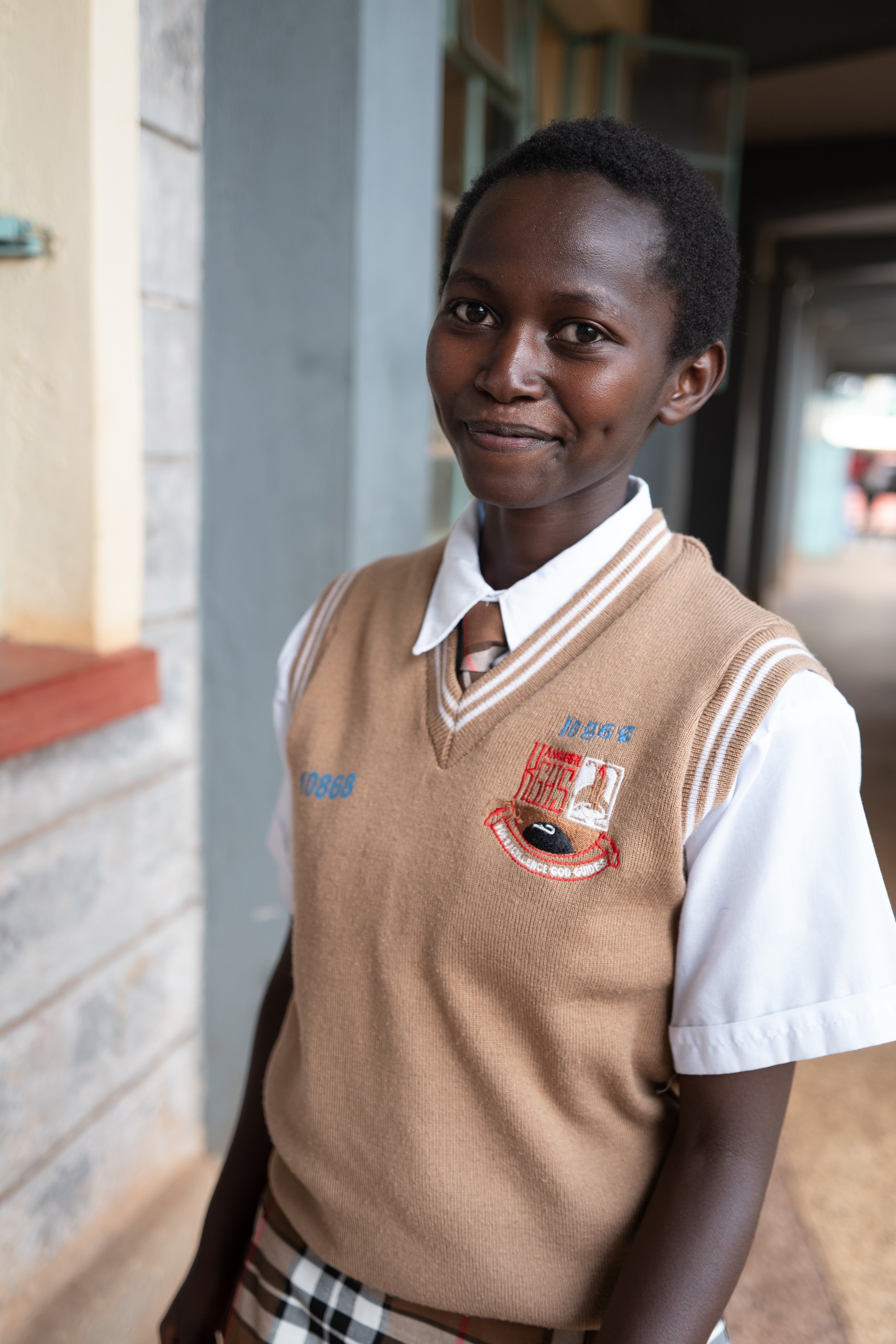 A girl in a school uniform standing in an outdoor corridor, smiling at the camera.