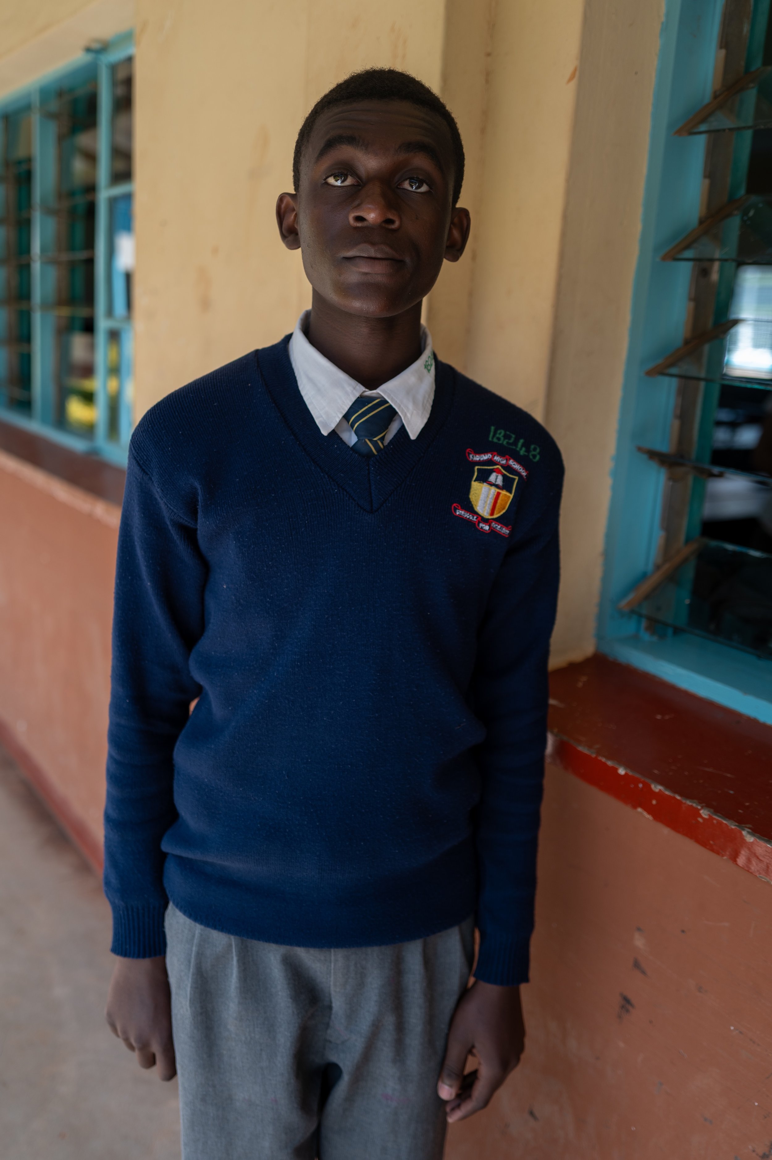A young boy in a school uniform standing in a classroom, looking up with a neutral expression.