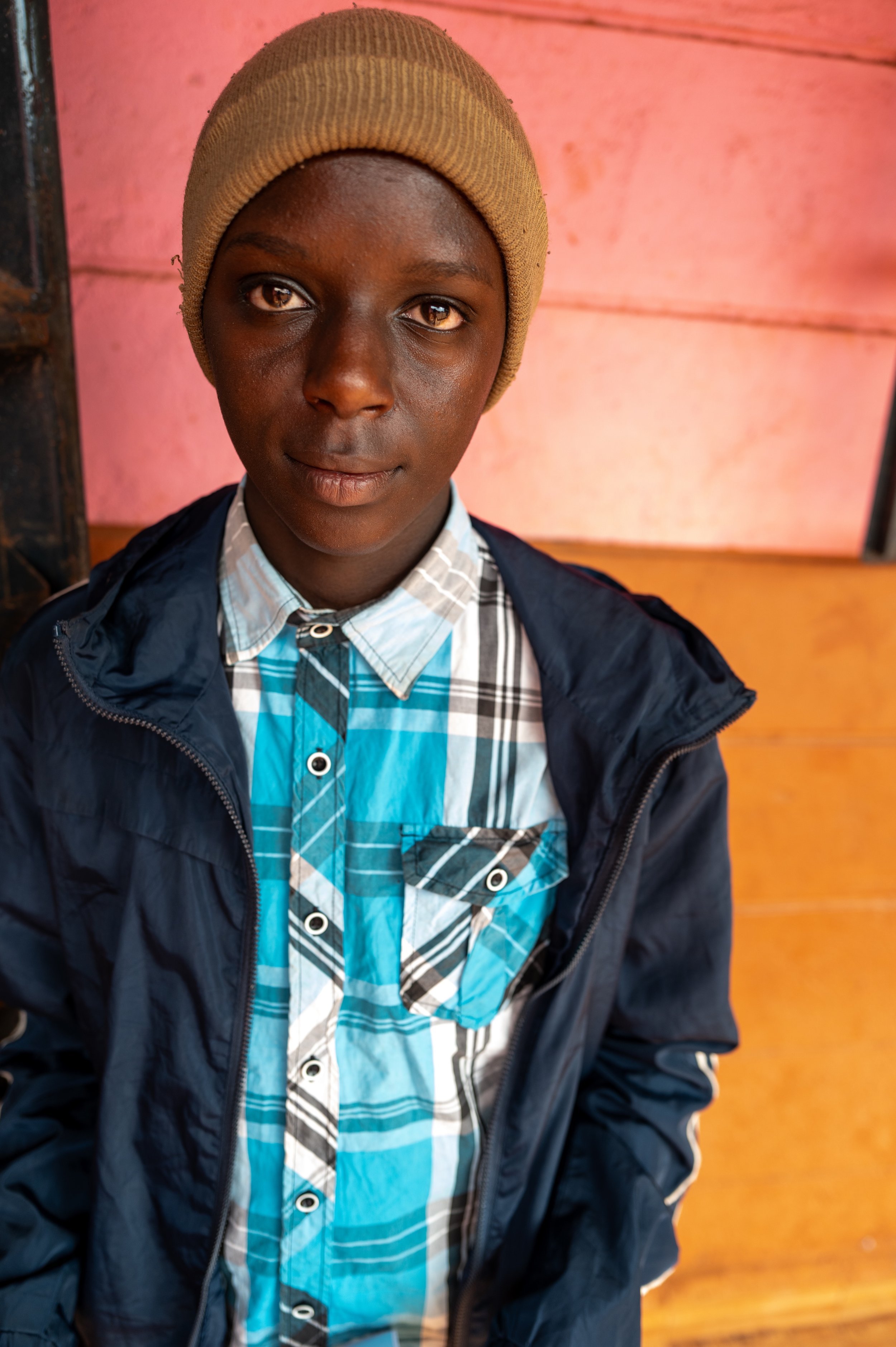 Young boy with dark skin wearing a beige knit beanie, a blue plaid shirt, and a dark jacket, standing in front of a pink wall and wooden background.