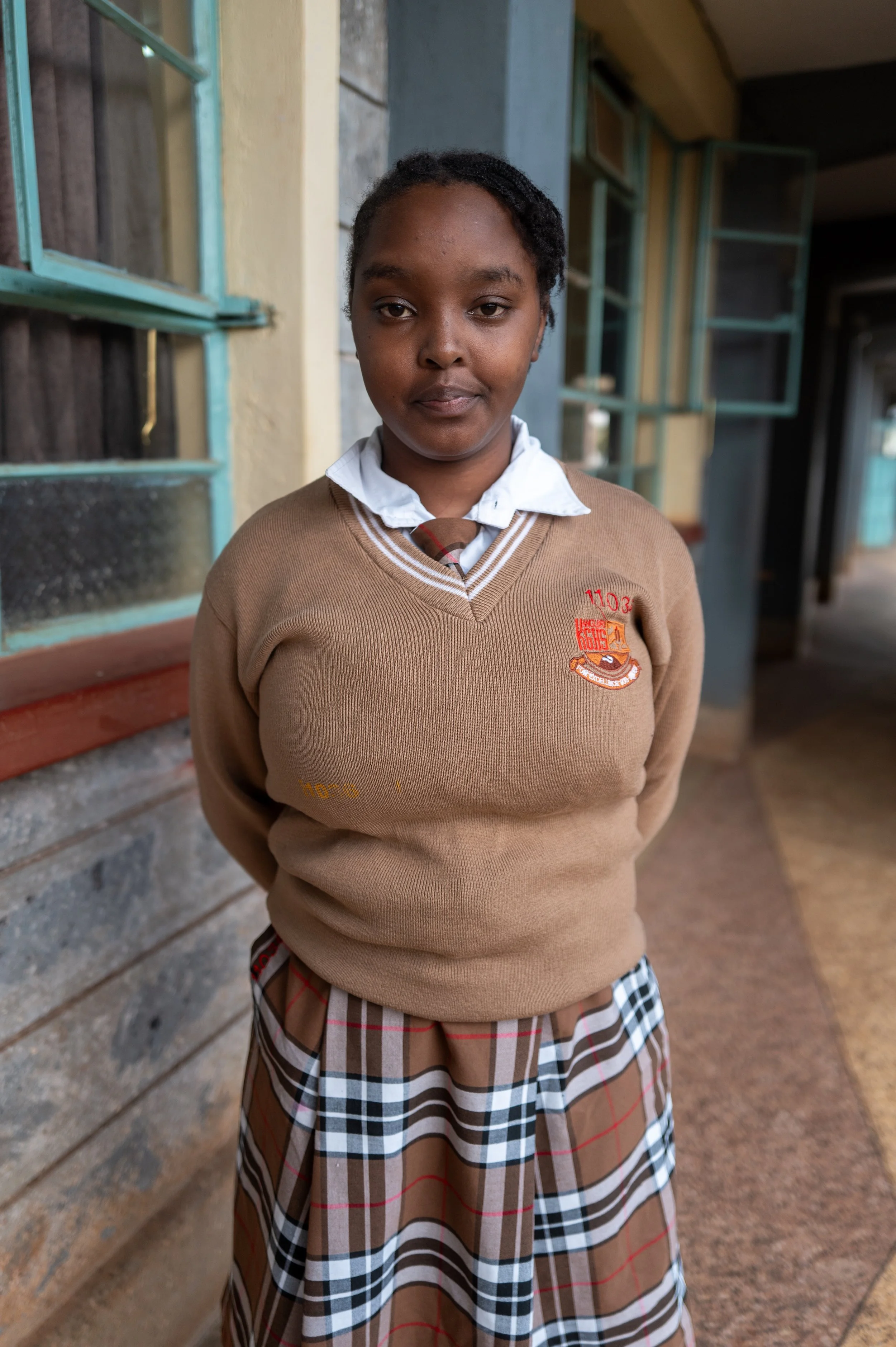 A young girl in school uniform standing outdoors near a building with windows and open shutters.