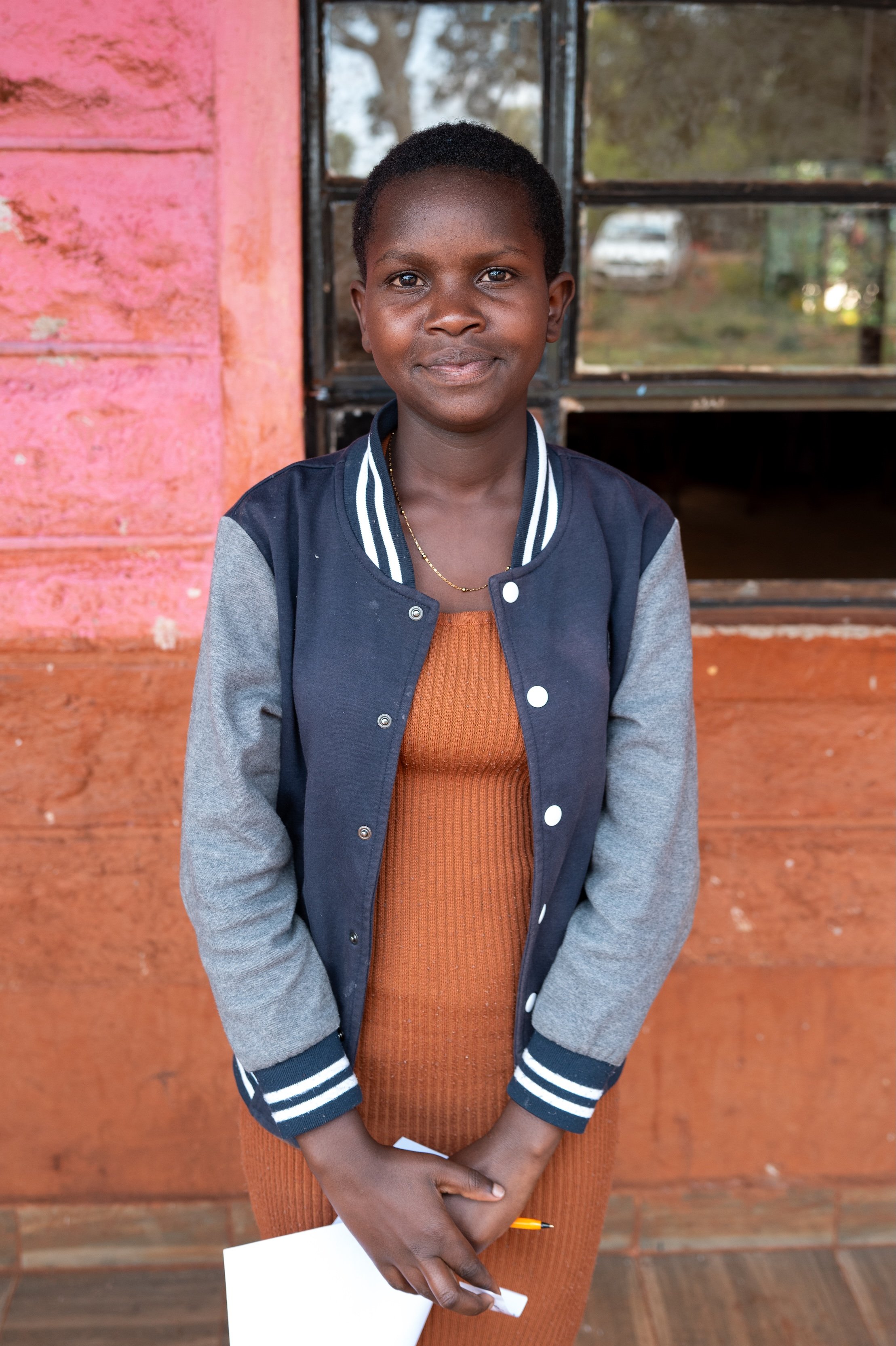 A young girl with short hair and dark skin standing outdoors in front of a pink wall and a window, wearing a navy blue and gray jacket over a brown dress, holding a white paper and a pen, with a slight smile.