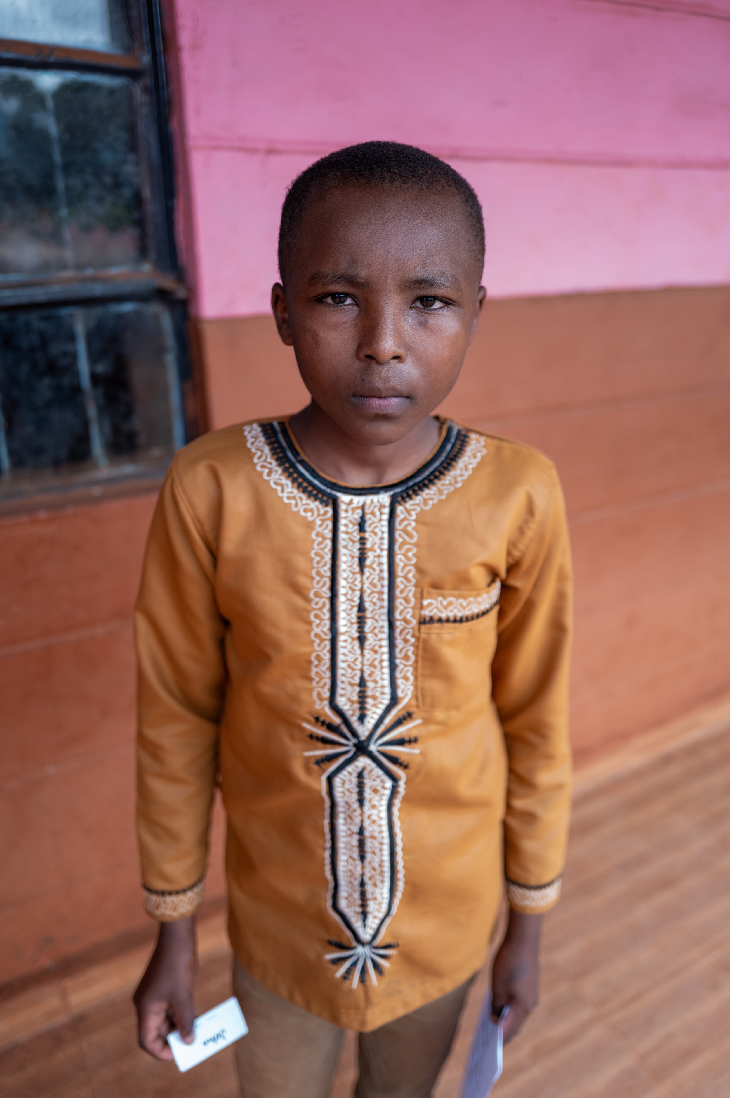 A young boy with short hair wearing a traditional orange-brown embroidered shirt, standing indoors in front of a pink and orange wall.