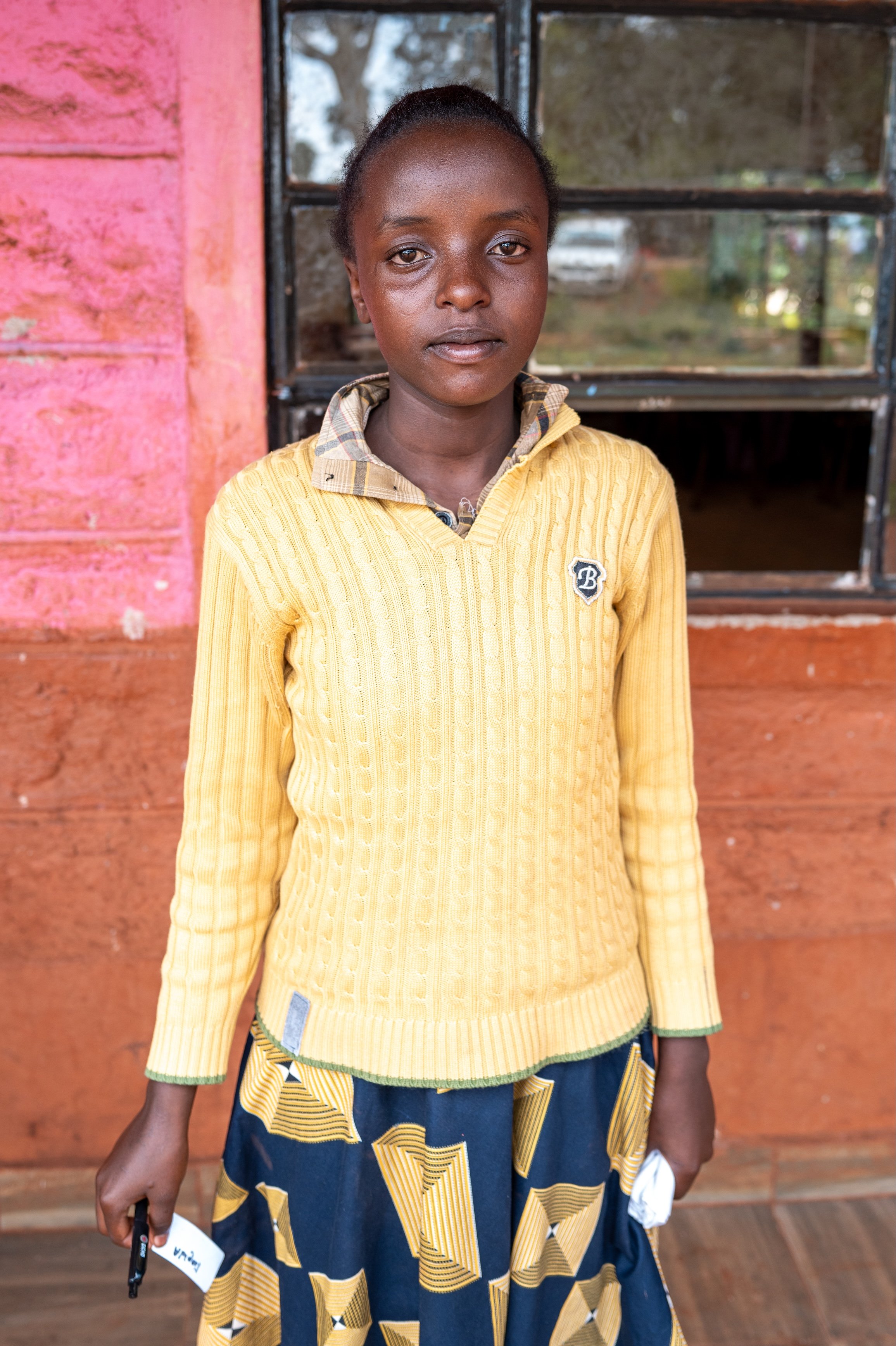 A young girl wearing a yellow sweater and a patterned skirt standing indoors in front of a window with a pink wall behind her.