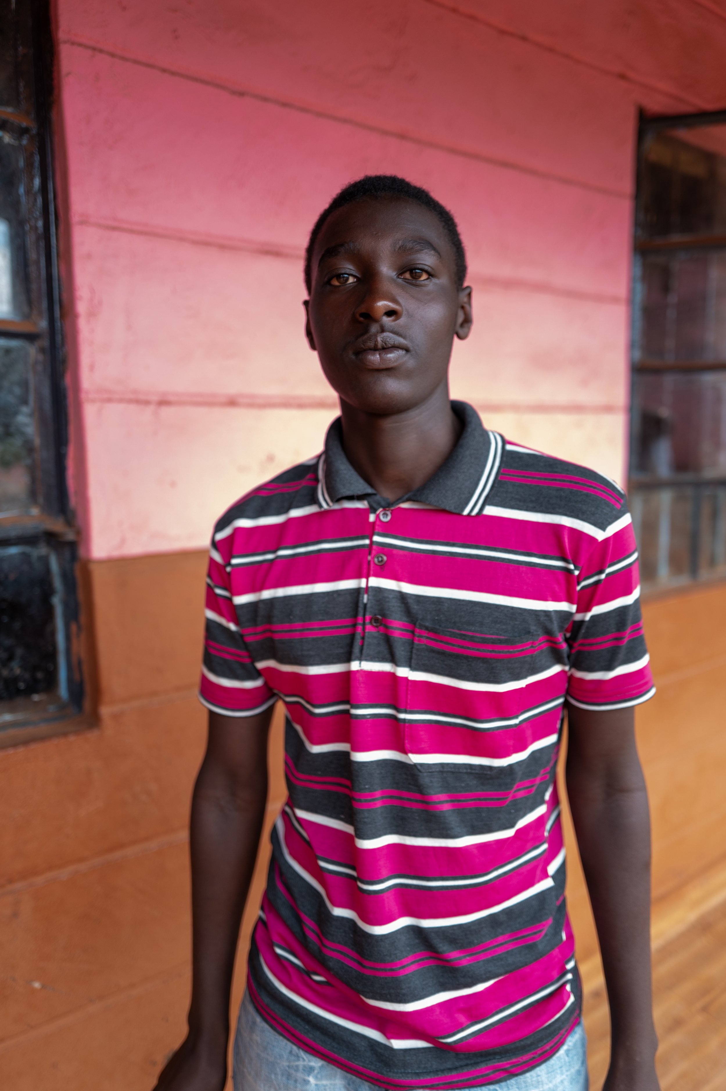 A young man standing in front of a pink and brown wooden wall, wearing a short-sleeved striped polo shirt.