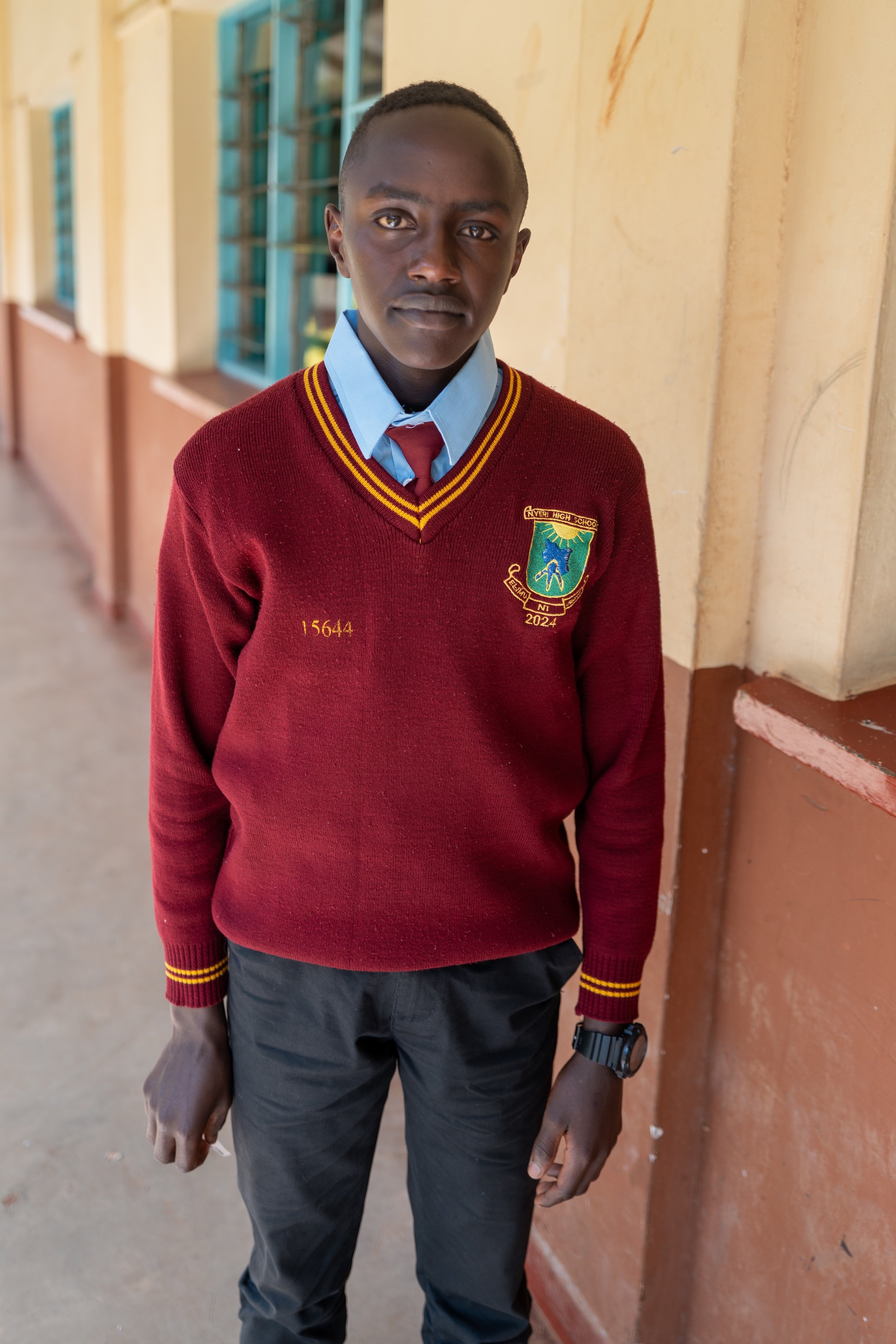 A schoolboy in a maroon sweater and blue shirt standing in a corridor of a school building.