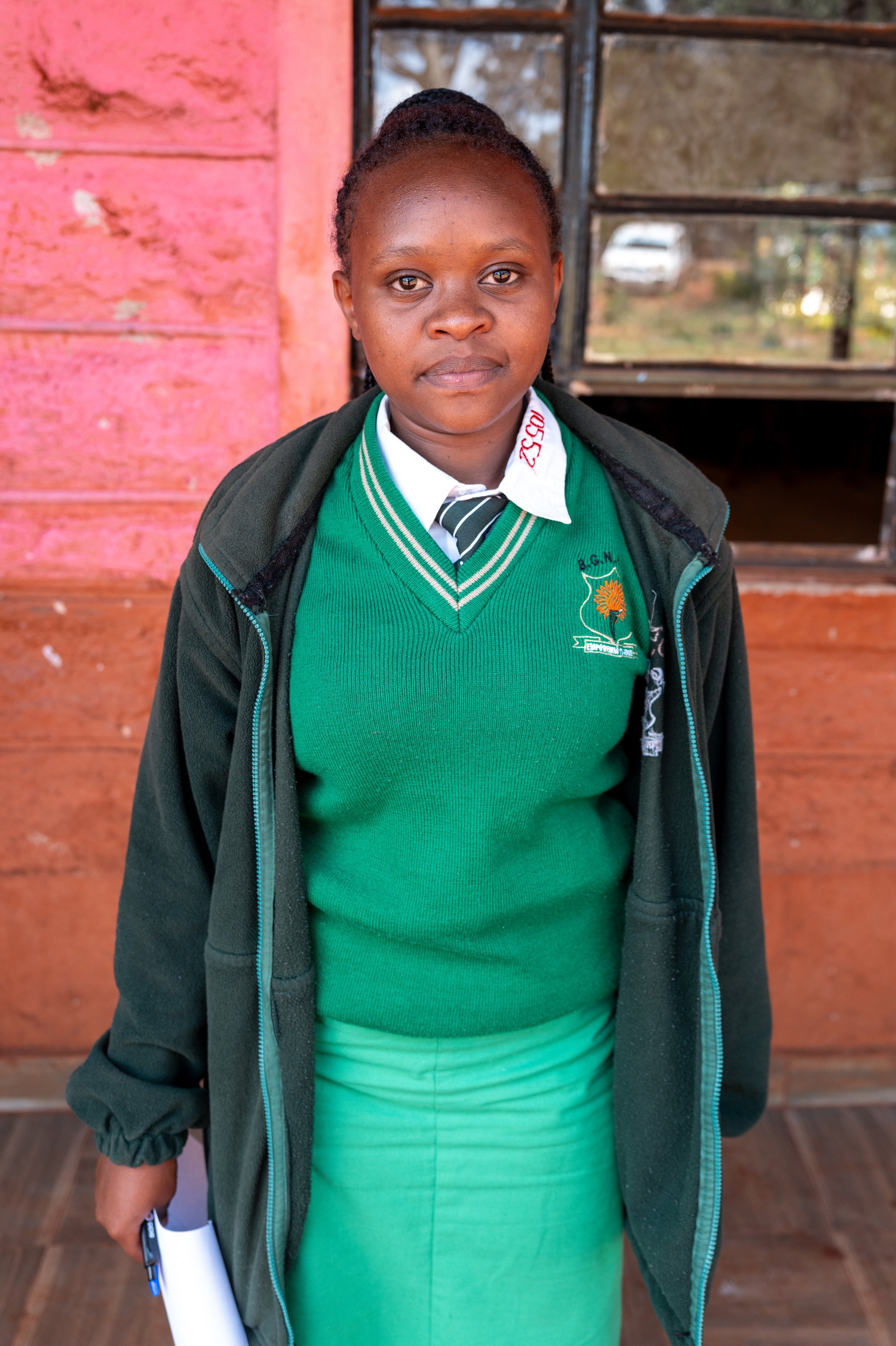A young girl in a school uniform standing outdoors in front of a wooden wall and window.
