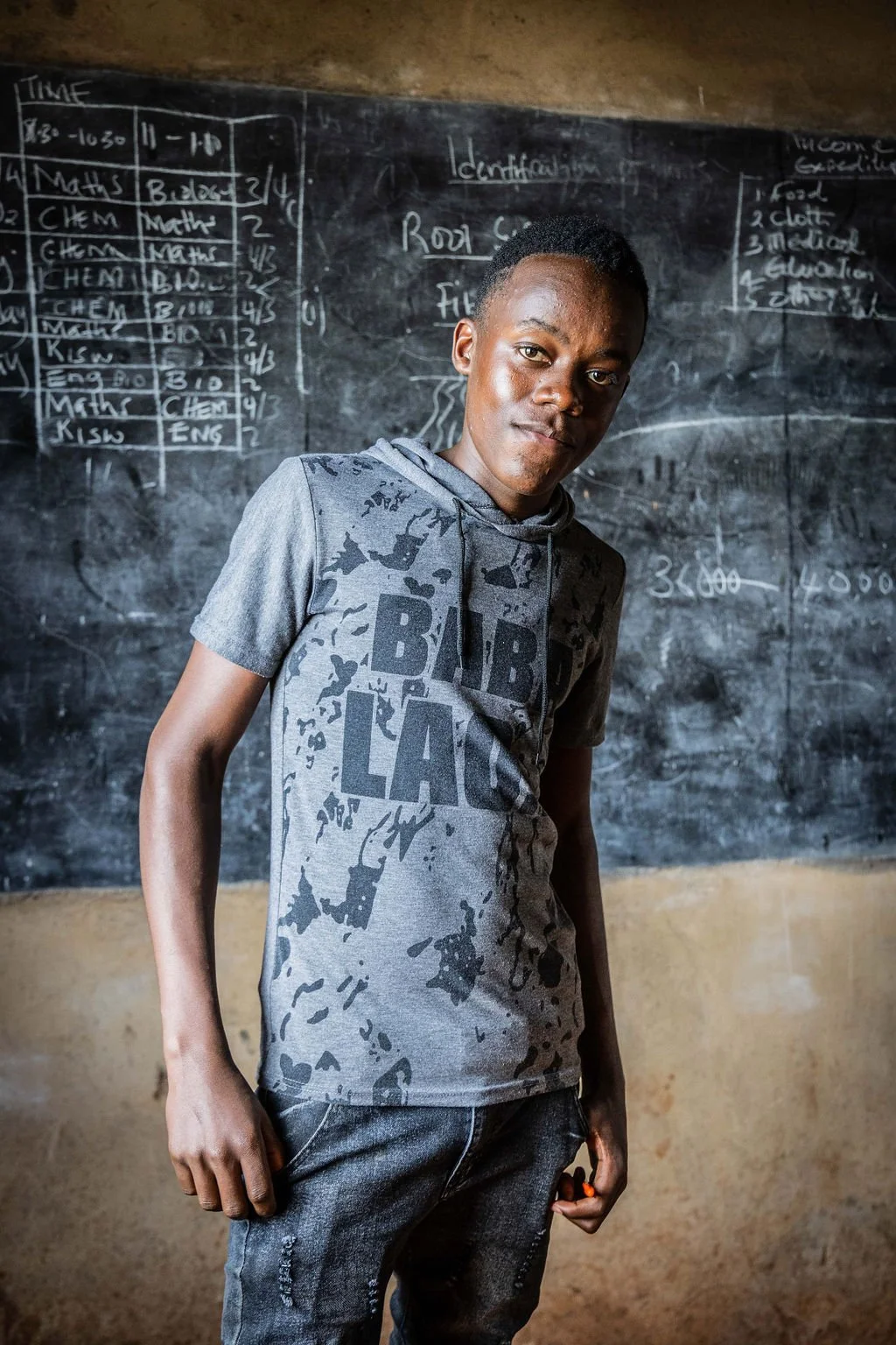 Young man standing in a classroom in front of a blackboard with writing and charts, wearing a gray graphic t-shirt.