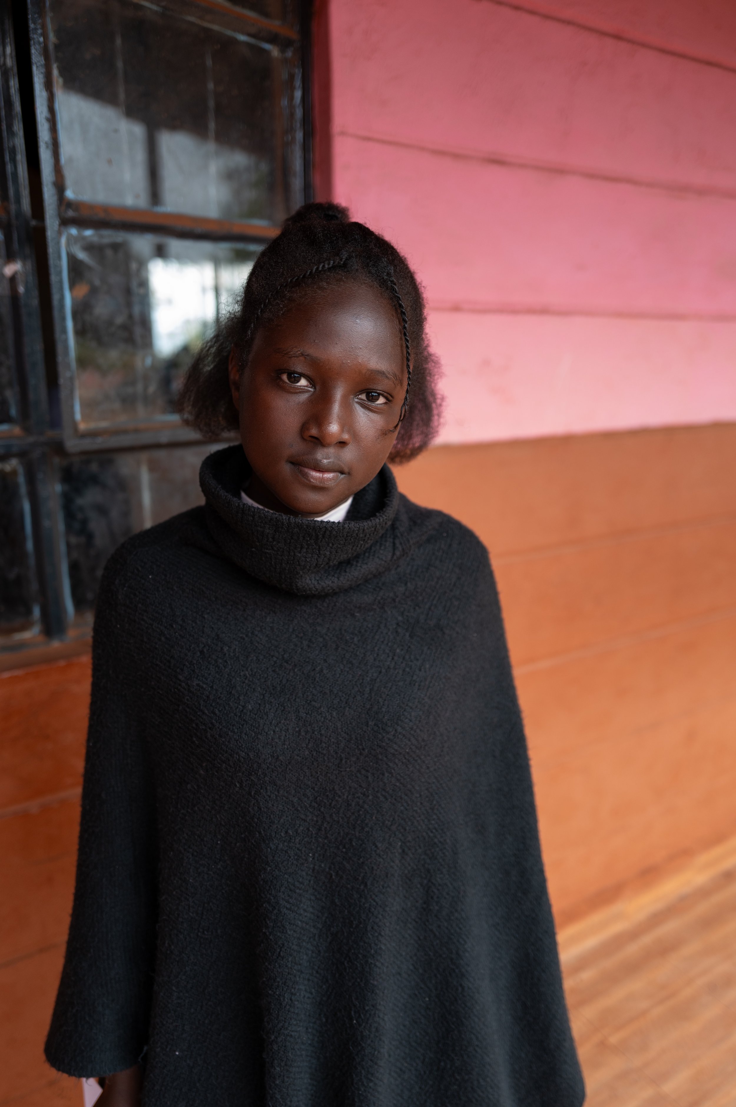 A young girl with dark skin and braided hair stands indoors in front of a colorful wooden wall and window. She is wearing a black turtleneck sweater and looks directly at the camera.